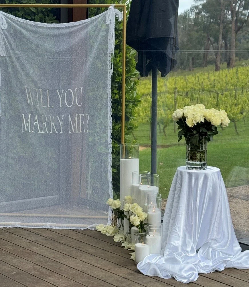 Romantic proposal backdrop with “Will You Marry Me” fabric signage, sand candles, and floral styling on Mornington Peninsula