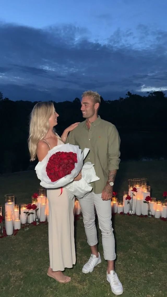 Outdoor proposal moment at dusk featuring candle styling, roses, and a couple celebrating together
