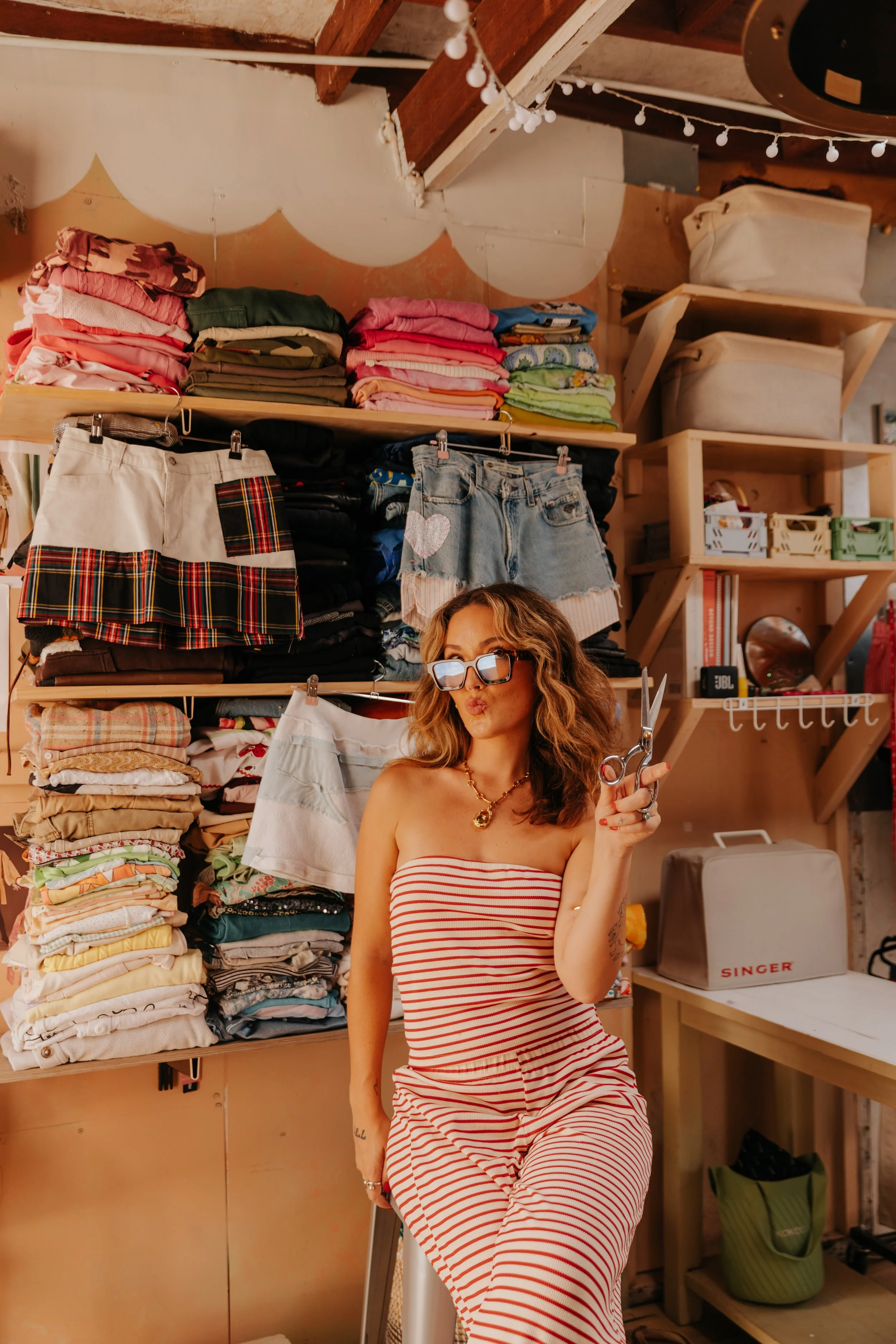 A woman with wavy hair, wearing sunglasses and a red-and-white striped strapless jumpsuit, holding scissors, standing in a room filled with folded clothes and storage shelves.