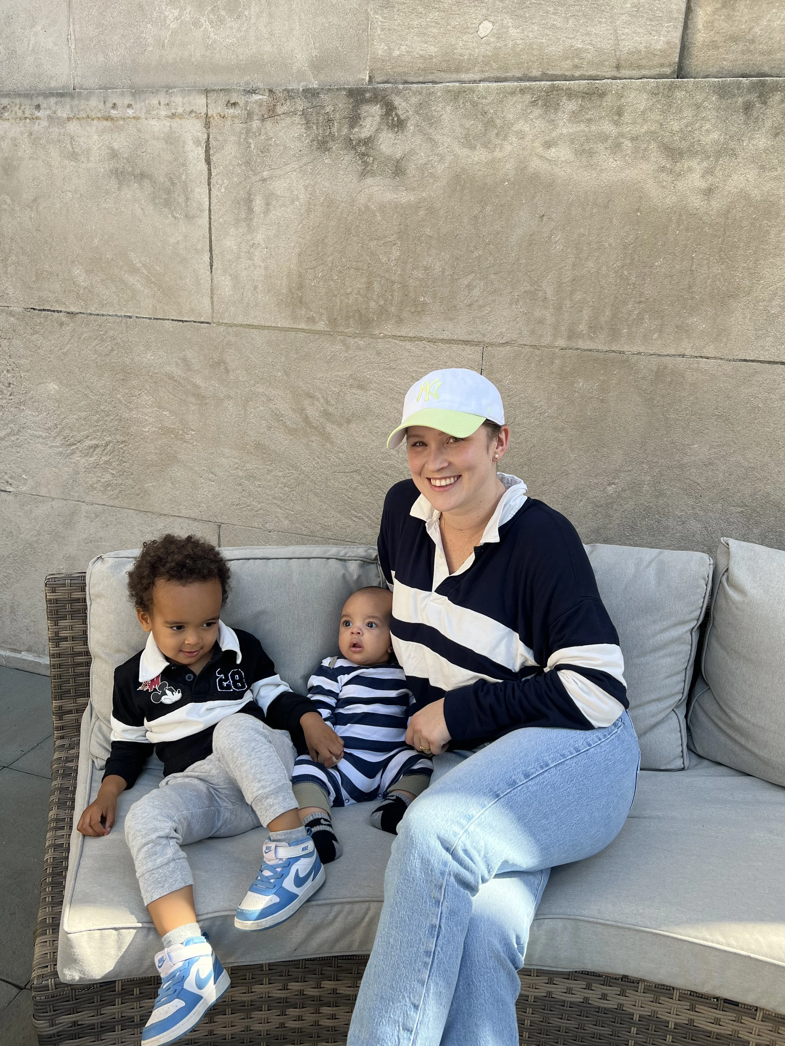 A woman sitting on an outdoor couch with two young children, all wearing striped and navy clothing. The woman has light skin, short hair, and is wearing a white cap and a black and white striped shirt. They are smiling and appear happy.