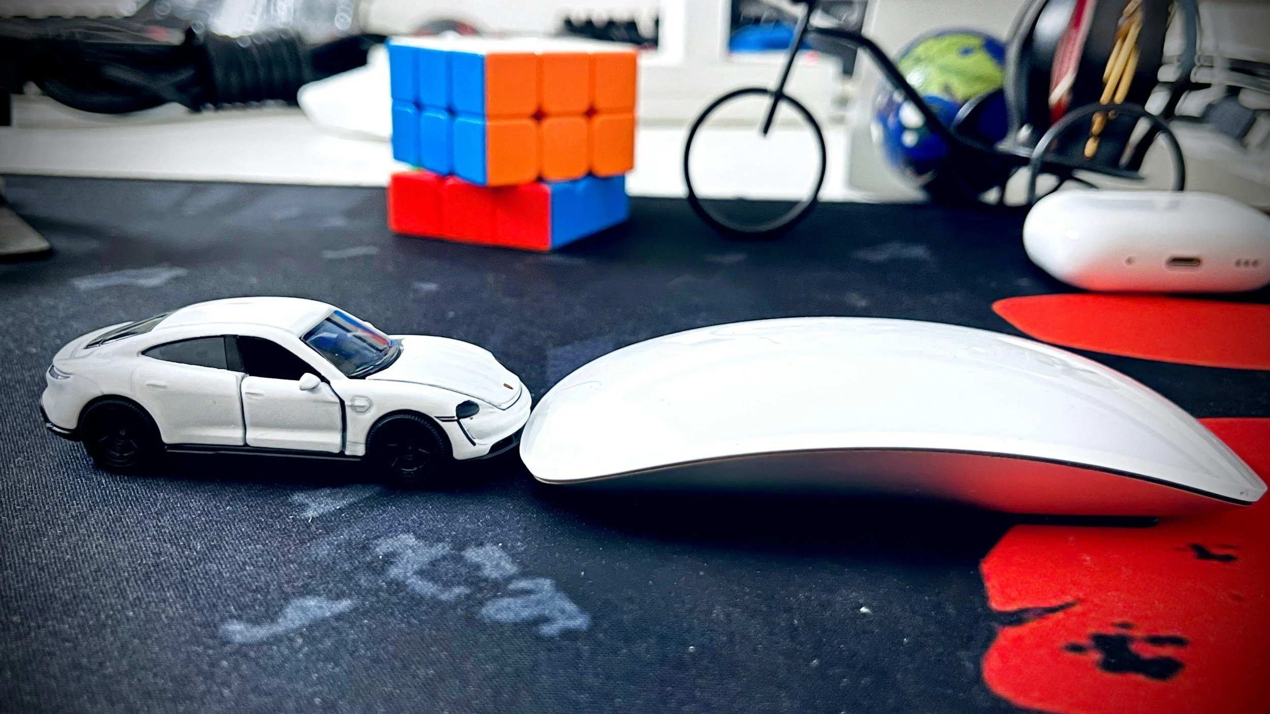 A small white toy car, a white computer mouse, a black-and-red keyboard wrist rest, a toaster, a black cube, and a bicycle-shaped keyholder on a black desk mat with red accents.