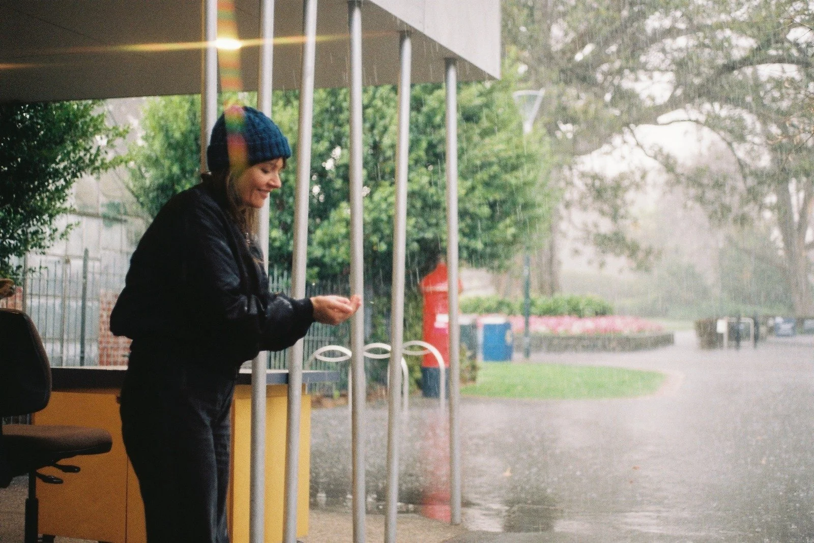 A woman standing inside a building near sliding glass doors, looking at her phone, wearing a blue knit hat and a black jacket, with rain outside and trees visible through the glass.