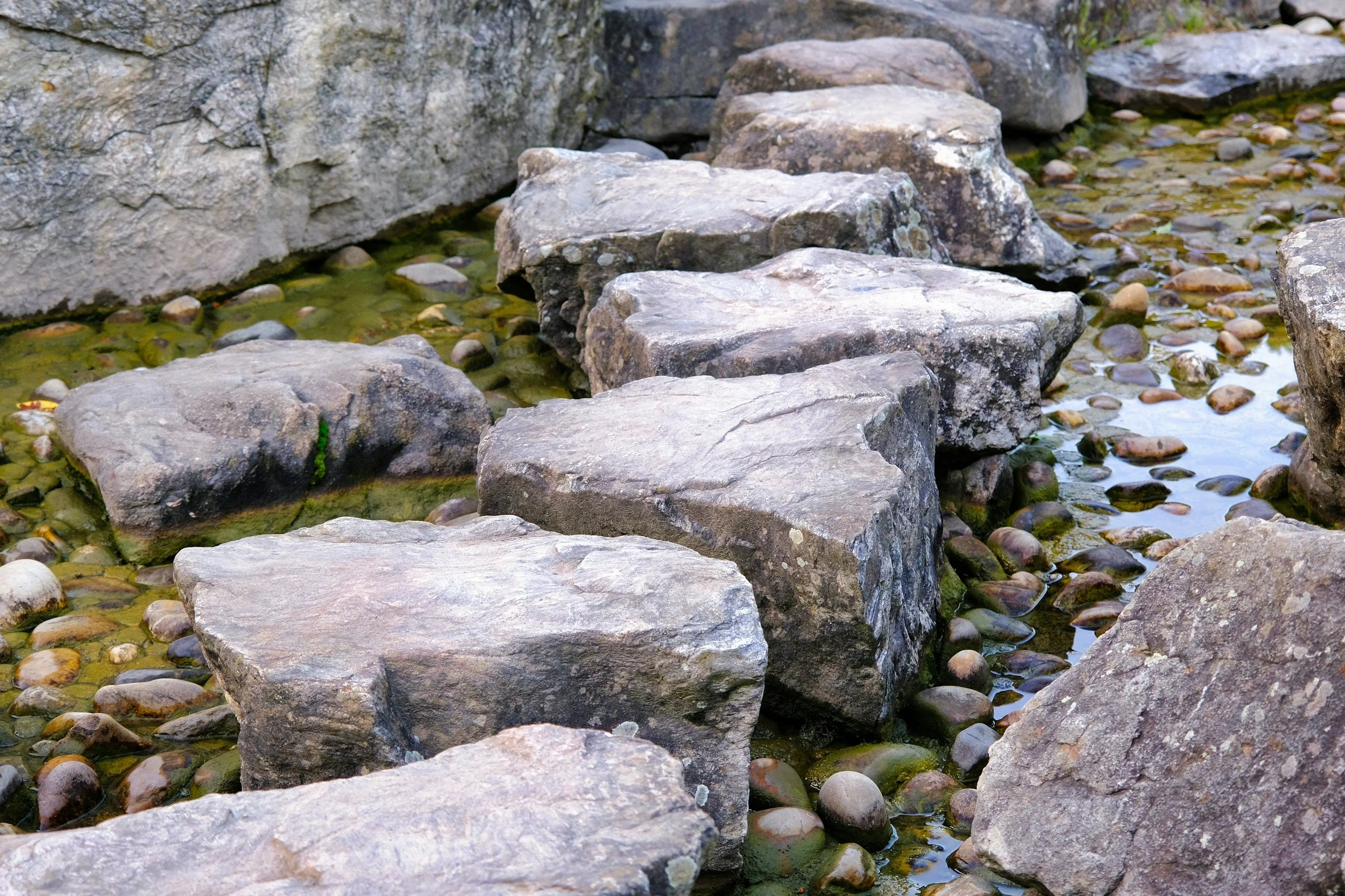 Large rocks and pebbles in a shallow stream of water with a mossy stone wall background.