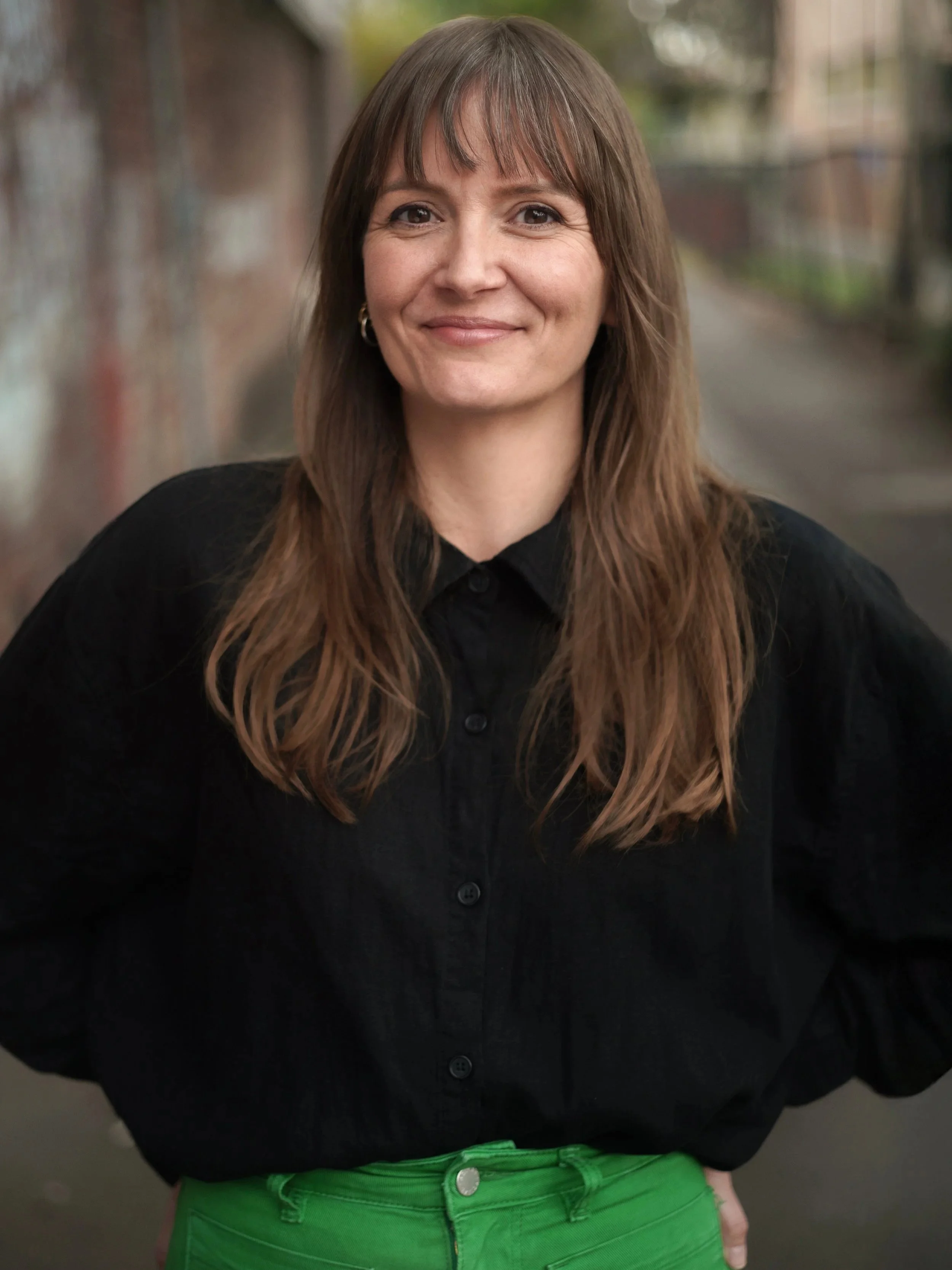 A woman with long brown hair, wearing a black button-up shirt and bright green pants, standing outdoors on a pathway with a blurred brick wall background.