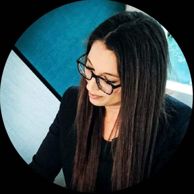A woman with long dark hair and glasses looking down, wearing a black blazer, sitting in a professional setting.