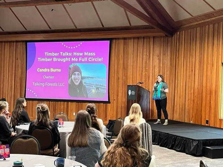 Candra Burns Speaking on the world Forestry Center Stage with black cloth and wooden walls near a projector with her past border collie dog spirit guide.