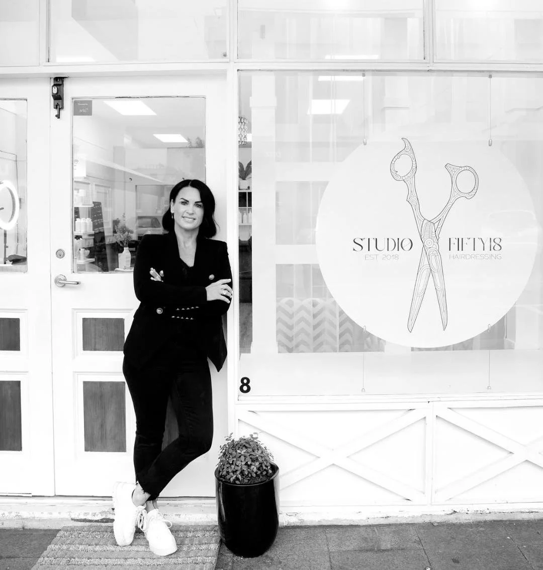 A woman with dark hair and a black blazer standing with arms crossed outside a hairdressing studio. The studio's sign features a pair of scissors and the text 'Studio Fifty18' with 'Est. 2018'.