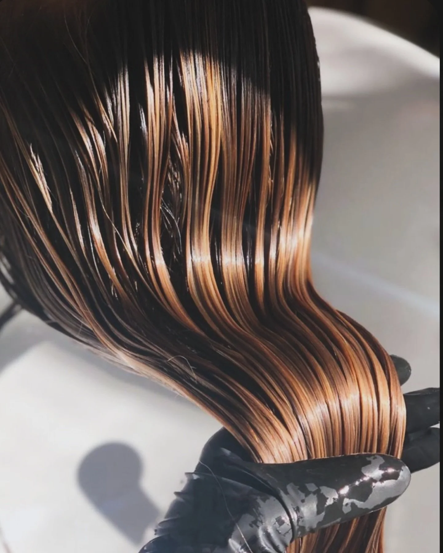 A person wearing black gloves holding a section of wet, styled hair with brown and black highlights.