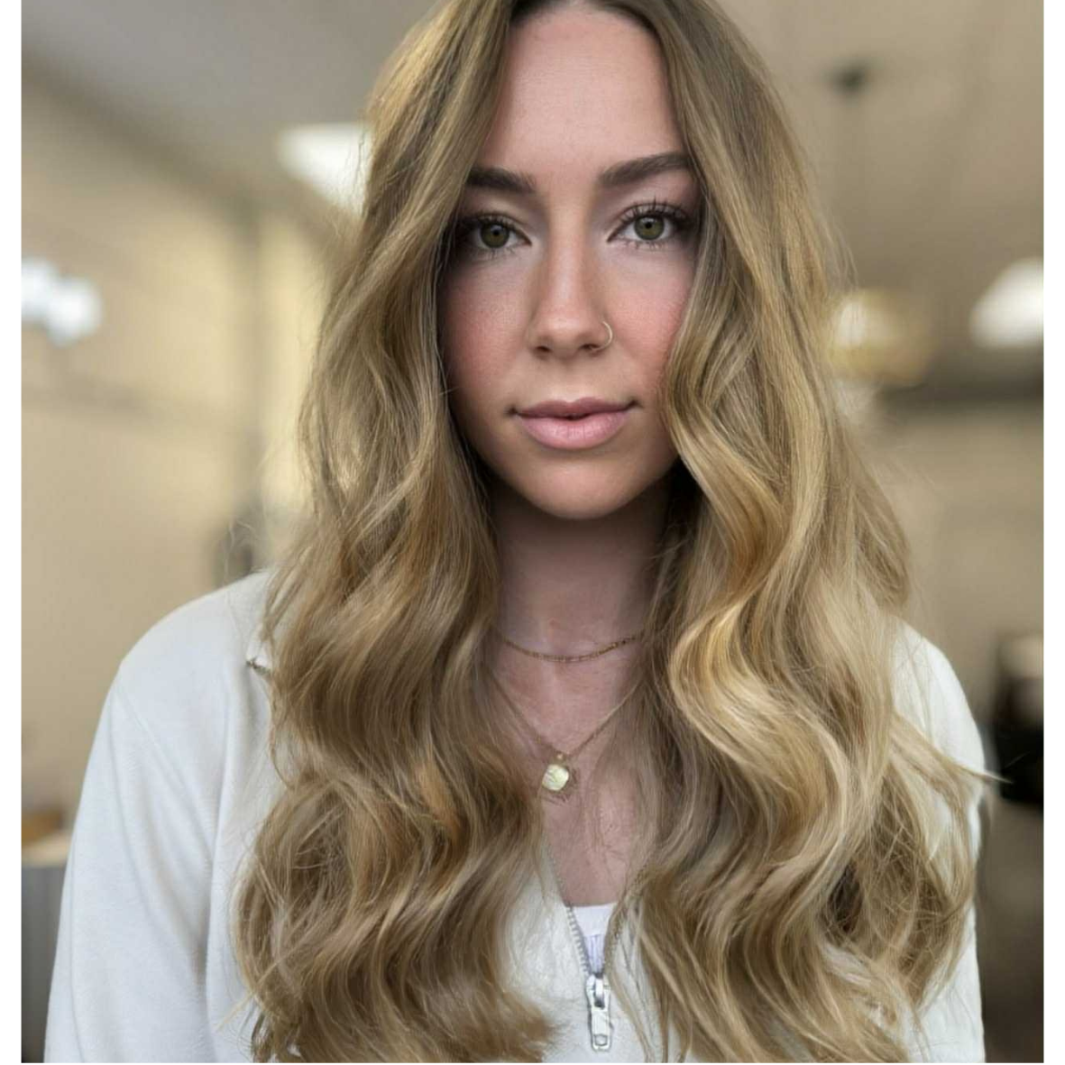 A woman with long wavy blond hair, light skin, wearing a white top and layered necklaces, looking into the camera indoors.