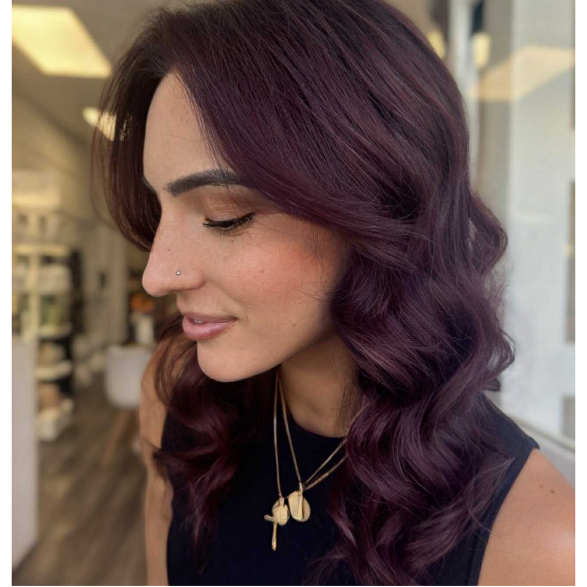 A woman with burgundy, wavy hair, closed eyes, a nose piercing, and layered necklaces, in an indoor setting.