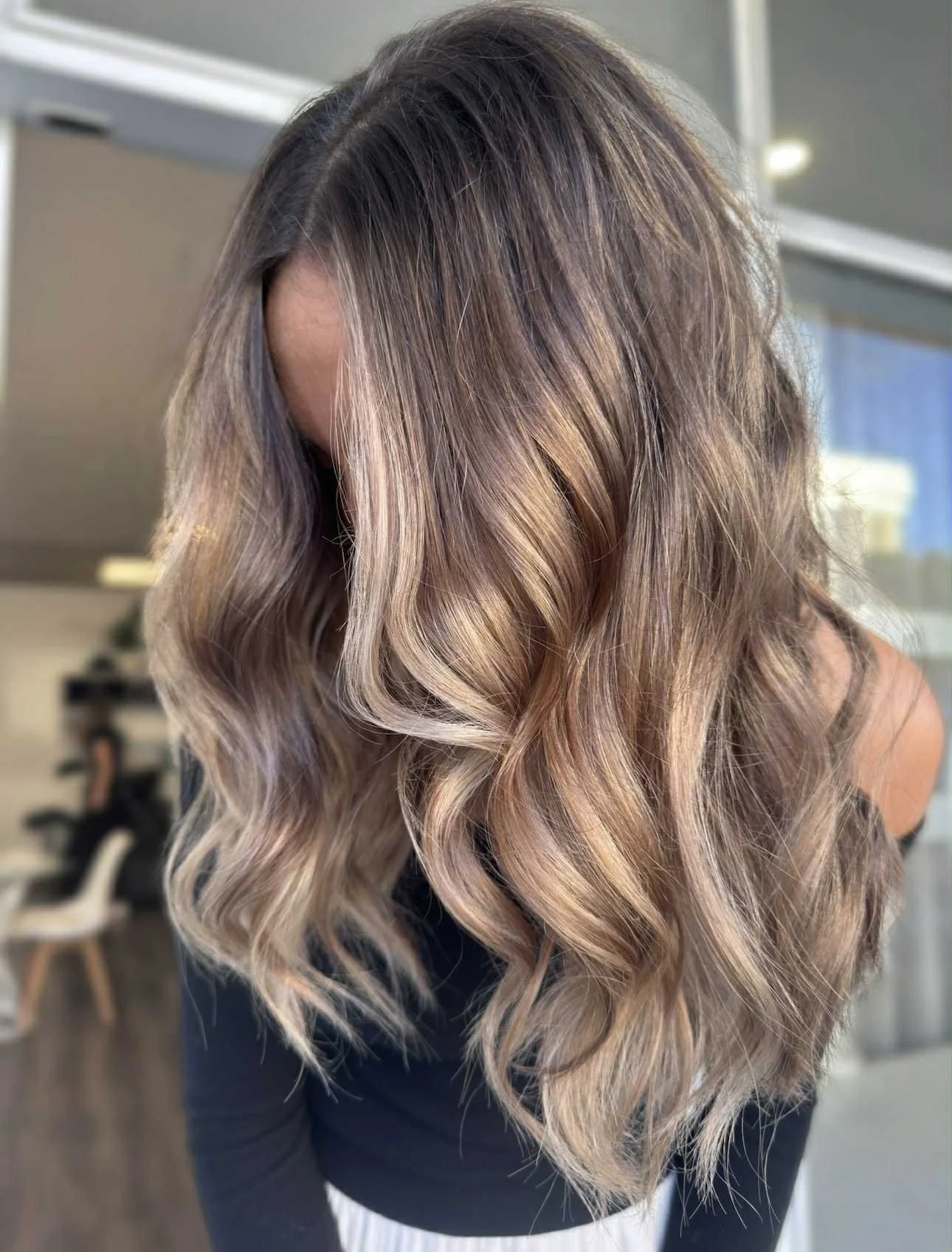 Close-up of a woman with long, wavy, ombre hair transitioning from brown at the roots to blonde at the ends, indoors with blurred background.