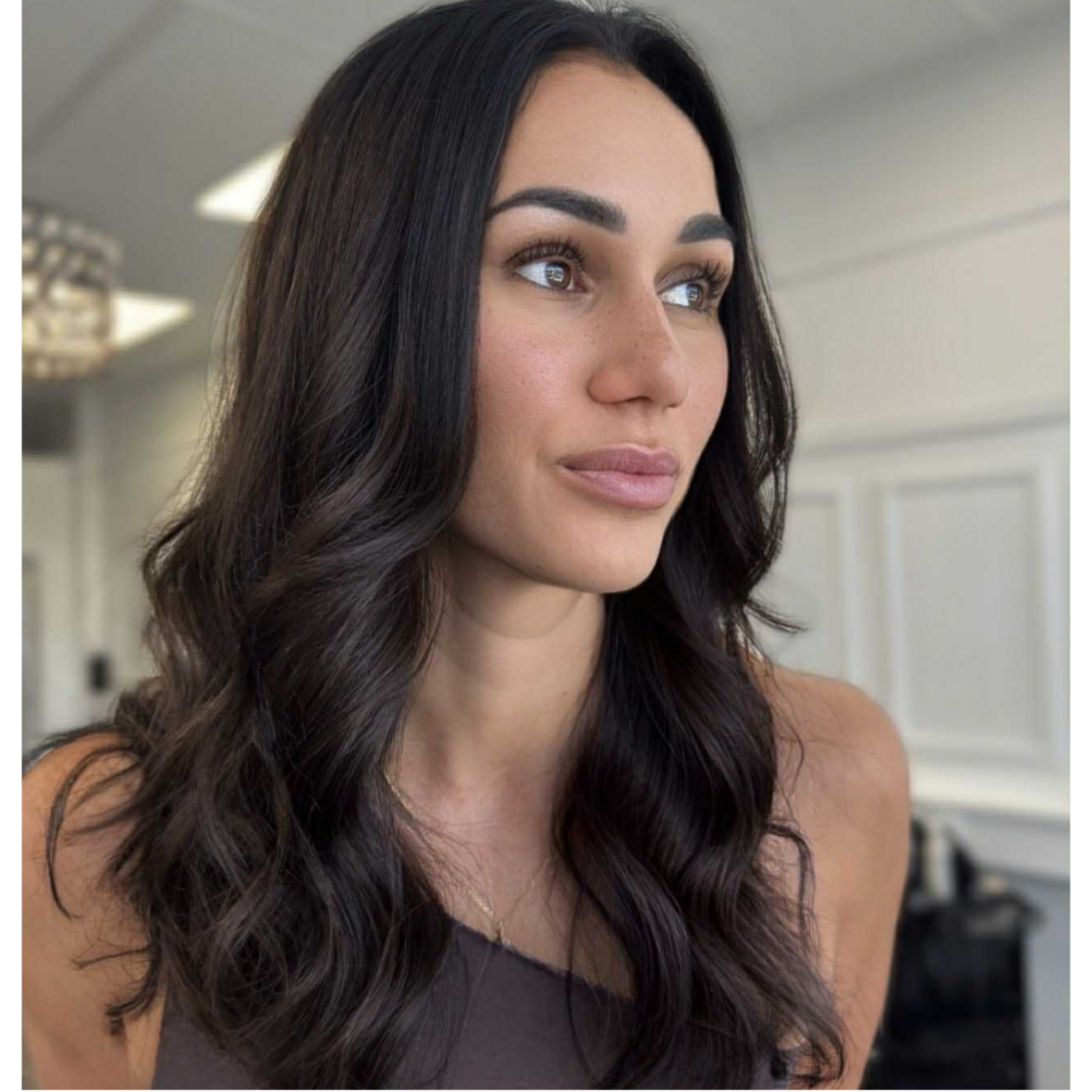 A woman with long, dark wavy hair looking to the side indoors.
