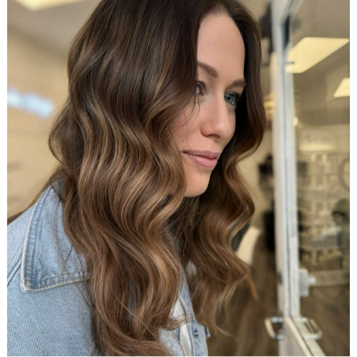 Close-up side profile of a woman with long wavy brown hair, blue eyes, and light skin, standing Near a glass window in a store or shopping mall.