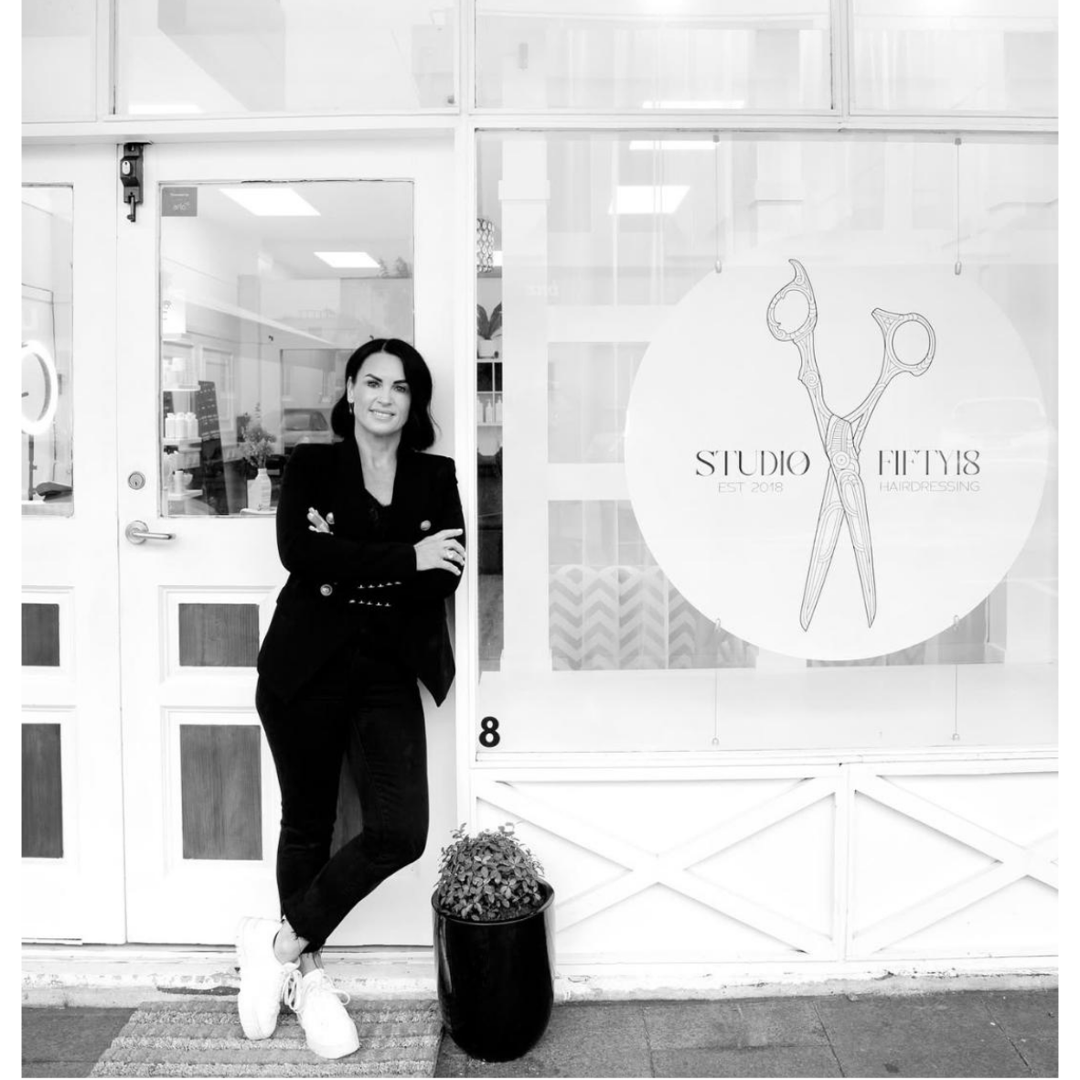 Woman standing outside hair salon with crossed arms, black outfit, and white sneakers, in front of a window with a salon logo featuring scissors, and a potted plant.
