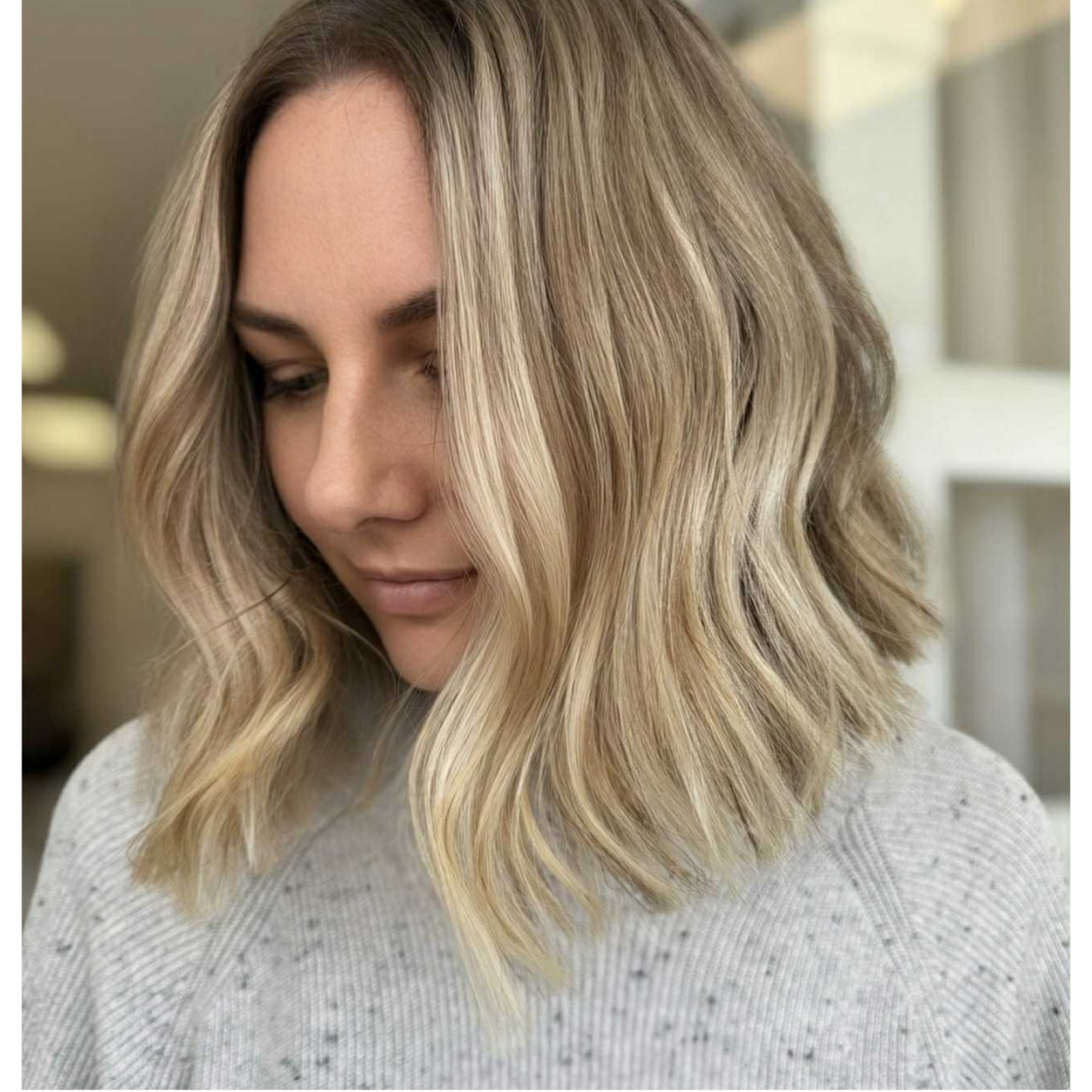 A woman with shoulder-length blonde wavy hair, wearing a light gray sweater, looking down gently.