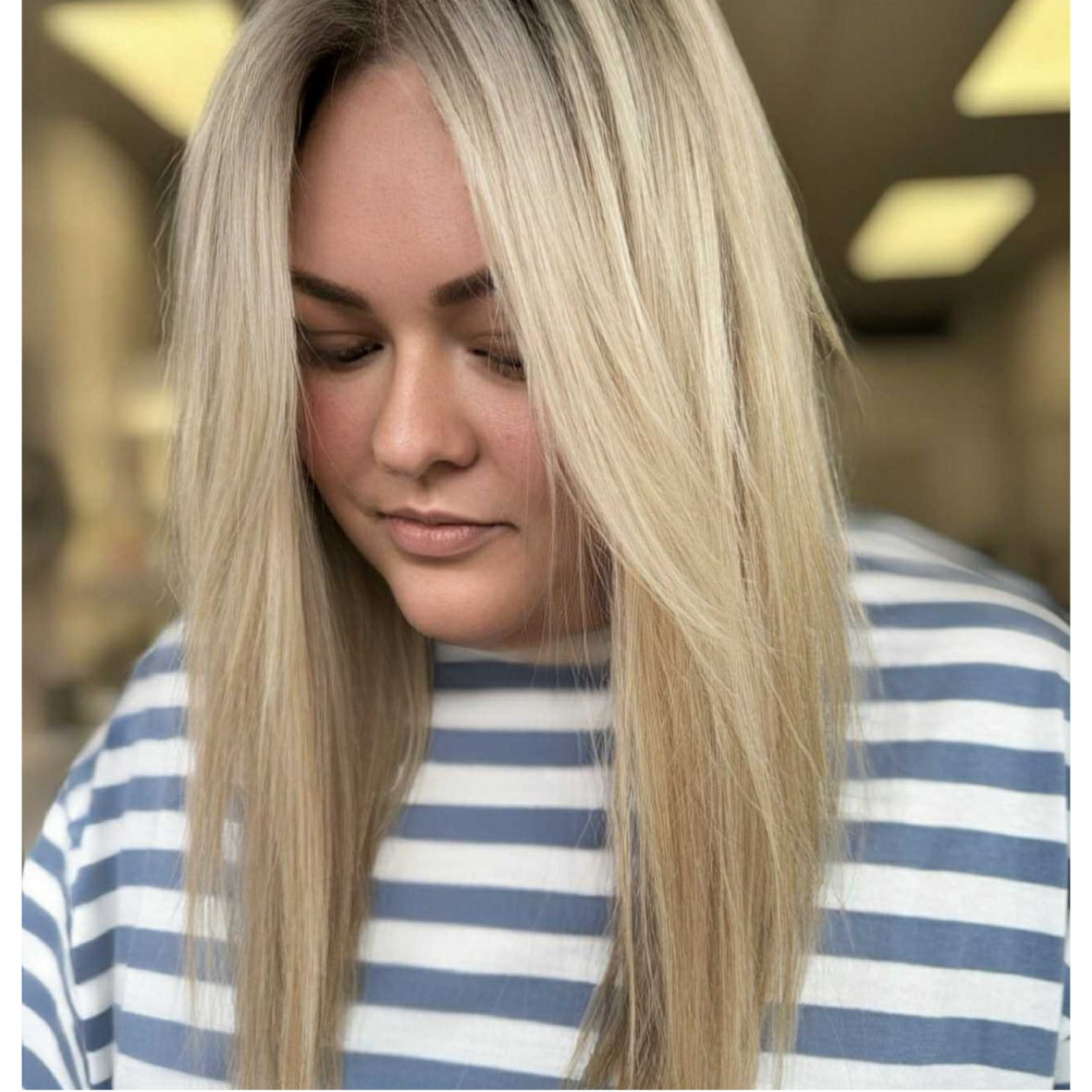 A young woman with long blonde hair wearing a blue and white striped shirt, looking down in a well-lit indoor space.