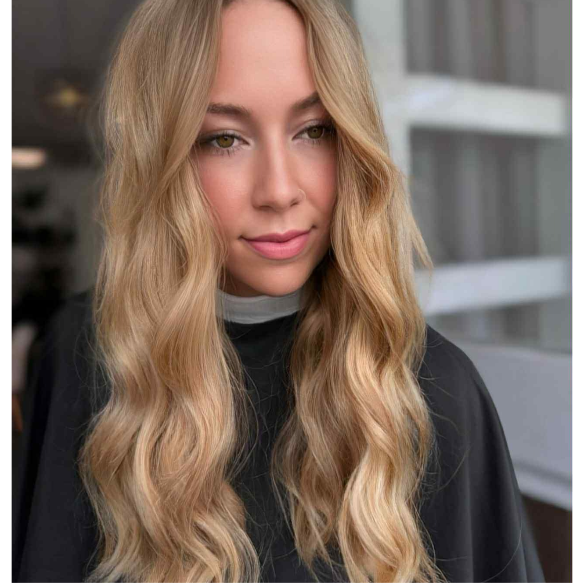 Close-up of a young woman with long, wavy blonde hair wearing a black top with a gray collar, standing indoors.