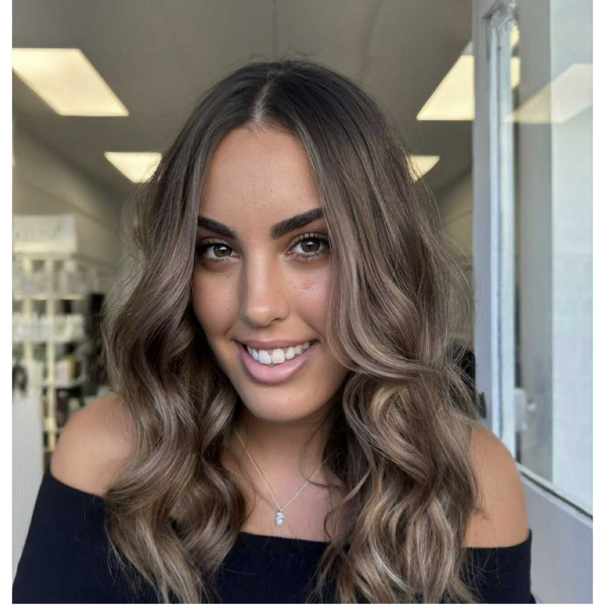 A young woman with wavy, shoulder-length hair and a bright smile, wearing a black off-shoulder top and a delicate necklace, indoors near a window.