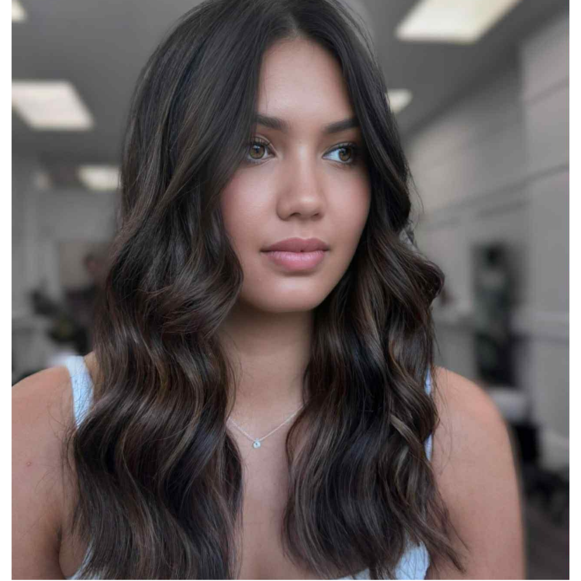 Close-up of a young woman with long, wavy brown hair and light makeup, wearing a light-colored top and a delicate necklace, in an indoor setting with a blurred background.