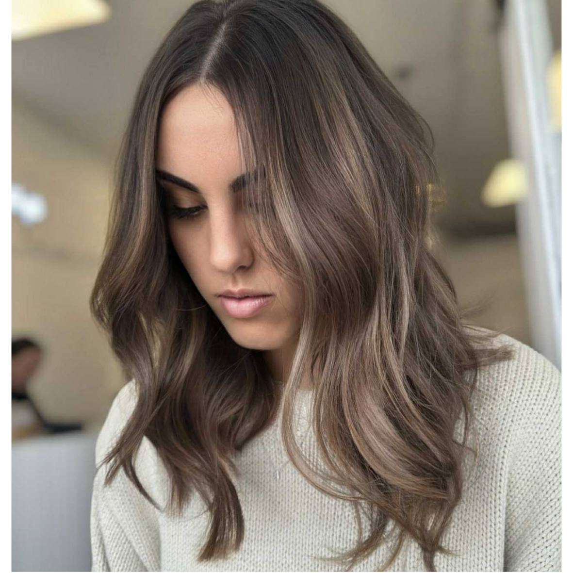 A woman with long, wavy brown hair looking down, wearing a cream-colored sweater, in an indoor setting.