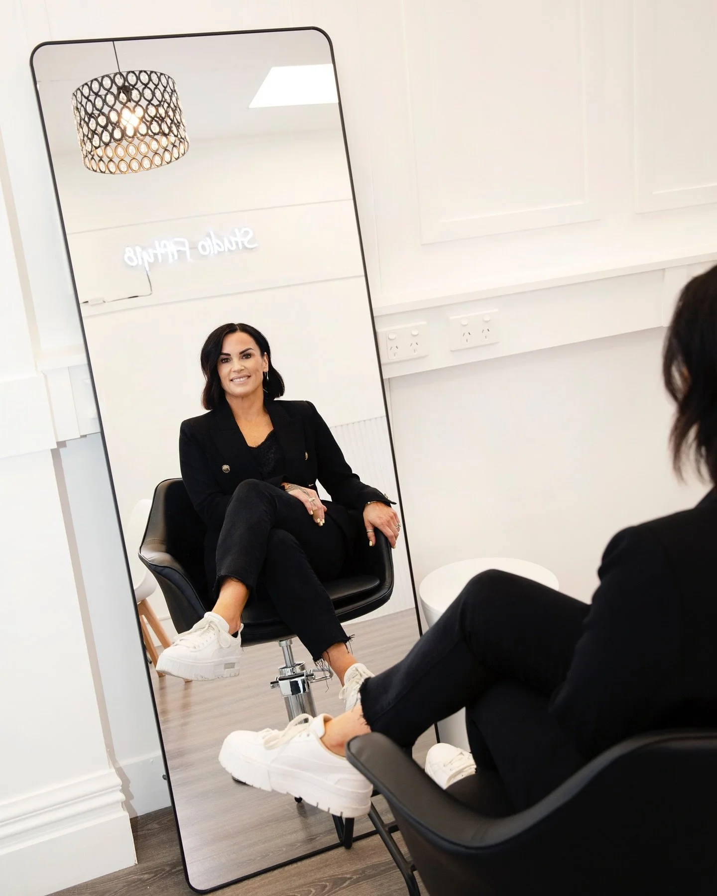 A woman in black clothing sitting in front of a large mirror, which reflects her smiling face and the word 'Studio' on a wall behind her.