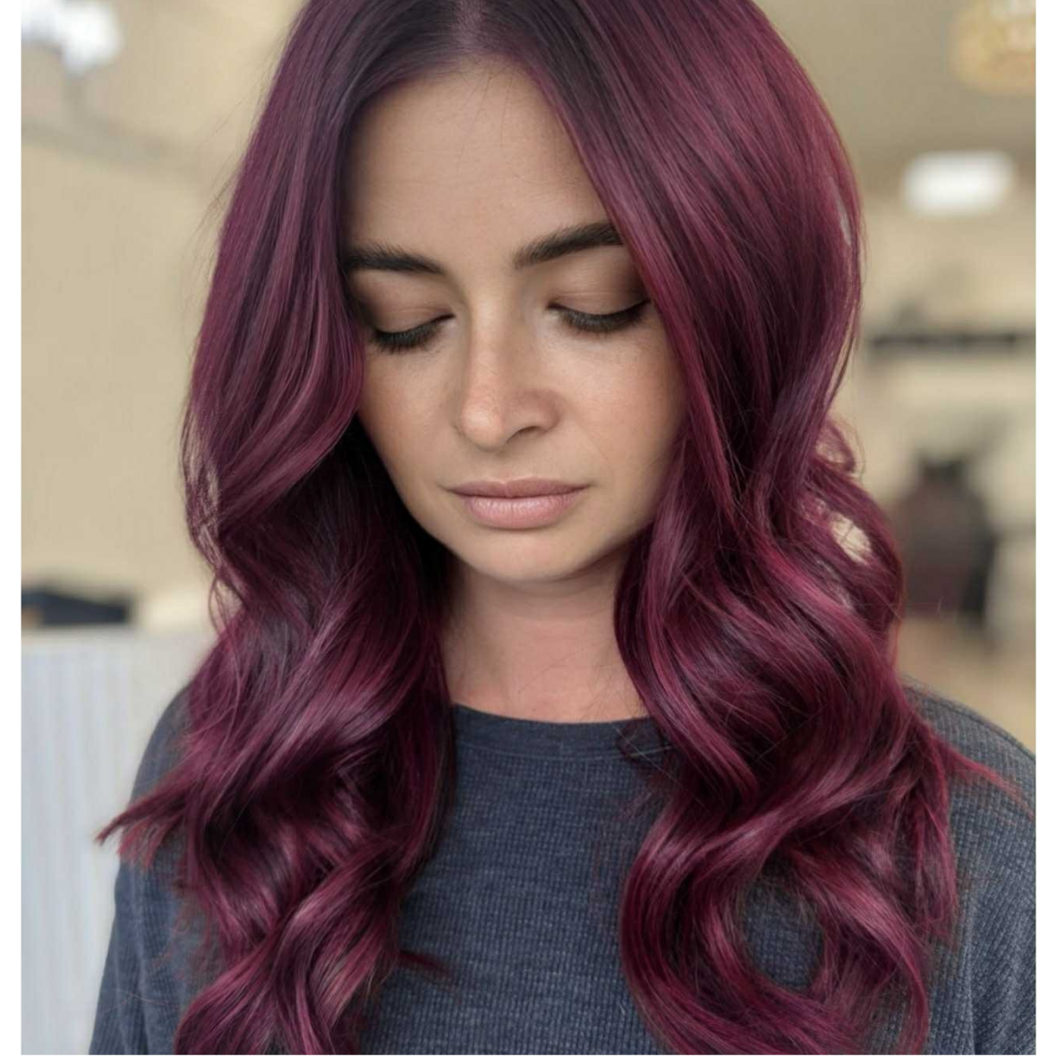 Young woman with long, wavy, magenta-colored hair, looking down with her eyes closed, in an indoor setting.