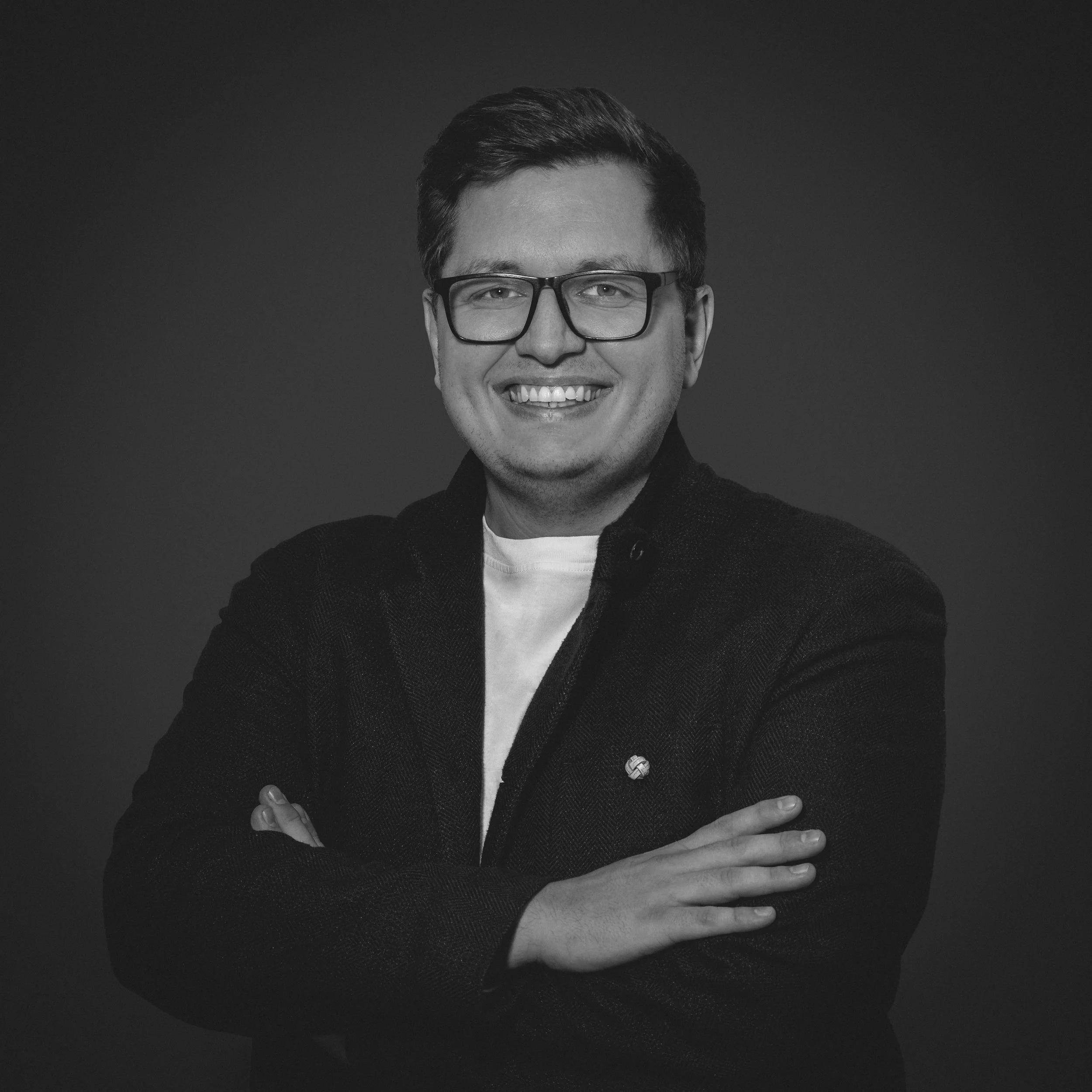 Jonas Hoettler mit Brille, lächelnd, mit verschränkten Armen, trägt einen dunklen Blazer und ein weißes T-Shirt, vor dunklem Hintergrund.