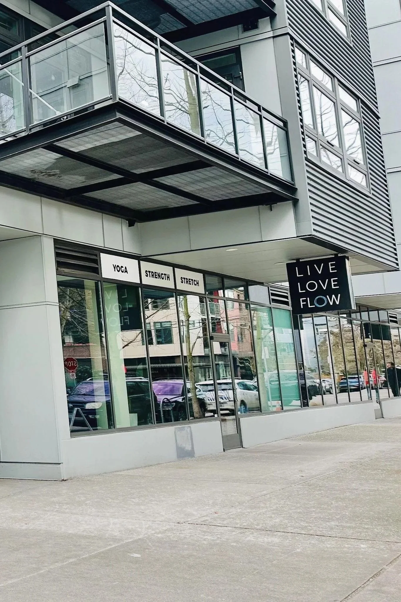 Exterior view of a modern fitness studio with large glass windows, signage indicating yoga, strength, and stretch classes, and a black sign hanging that says 'Live Love Flow.'