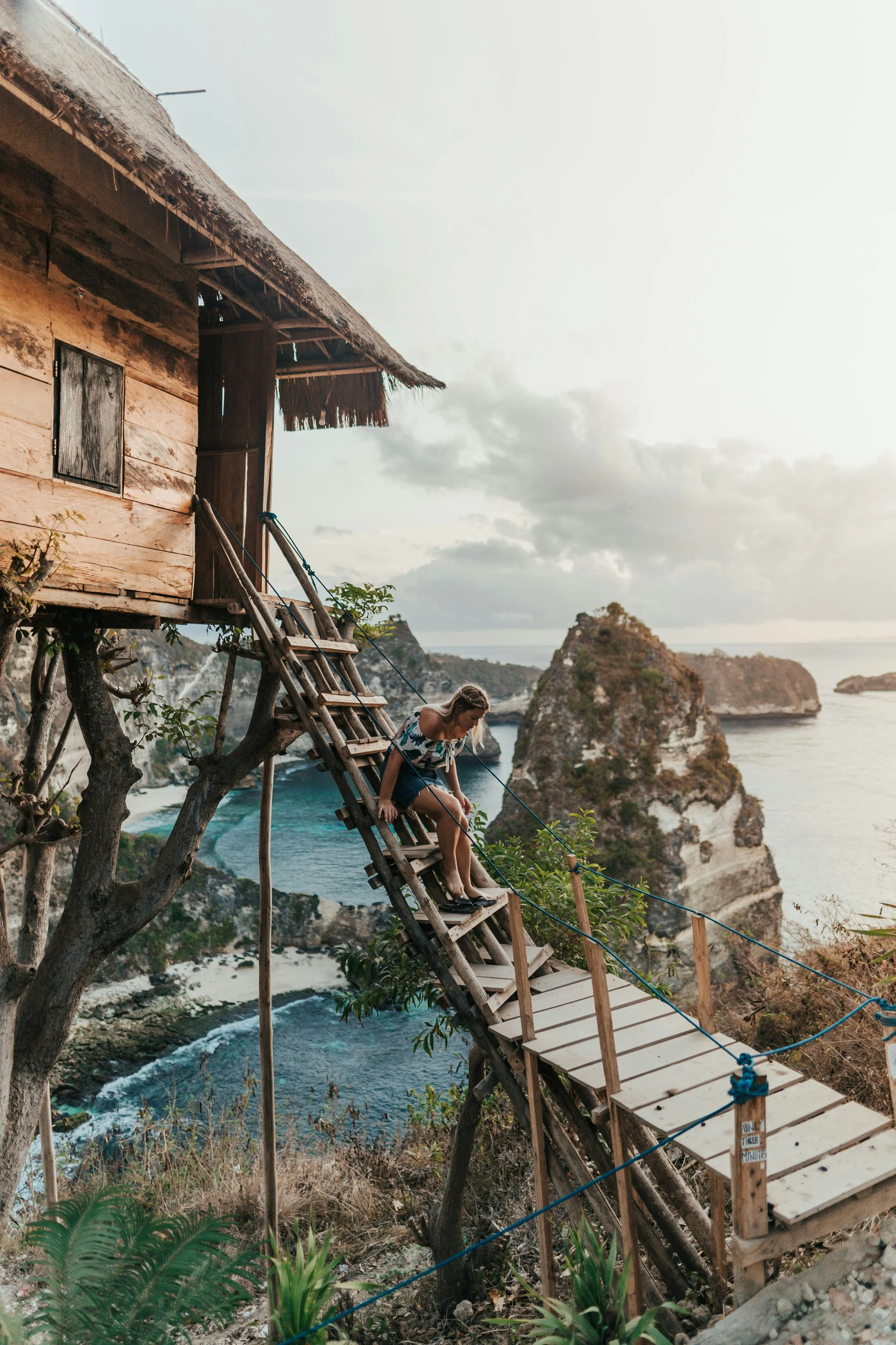 Person descending wooden stairs from a treehouse overlooking an oceanfront cliff landscape with turquoise waters.