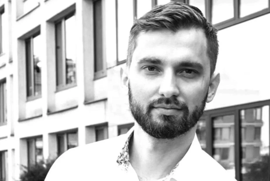 Black and white portrait of a young man with short hair and a beard, standing outdoors near a modern building with large windows.