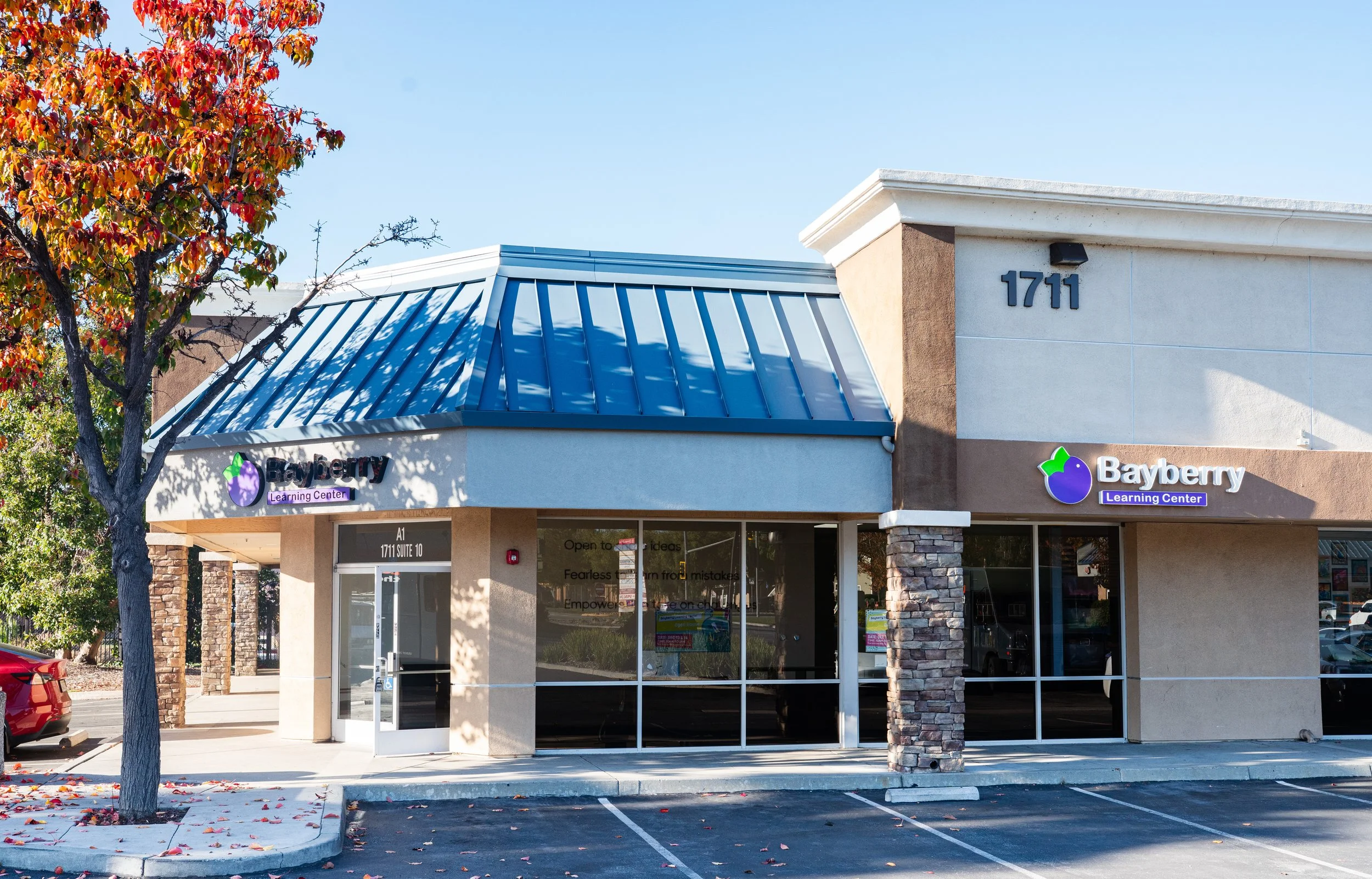 Exterior view of Bayberry Learning Center with a blue metal roof, a tree with red leaves, and parking spaces in front.
