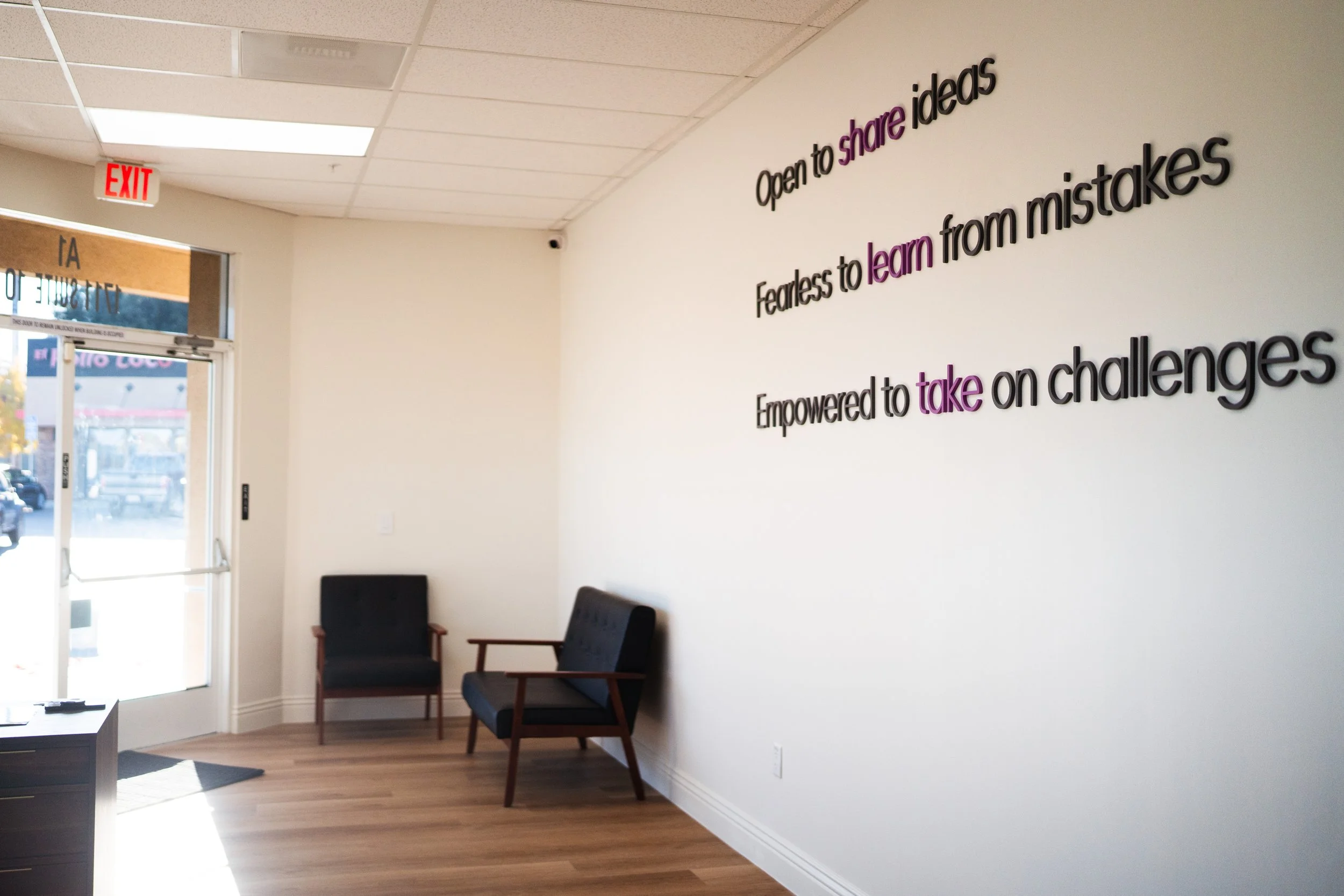 Interior of a waiting room with a white wall that has motivational quotes in black and purple text, two black chairs with wooden arms, a small black table near the glass door, and an exit sign above the door.
