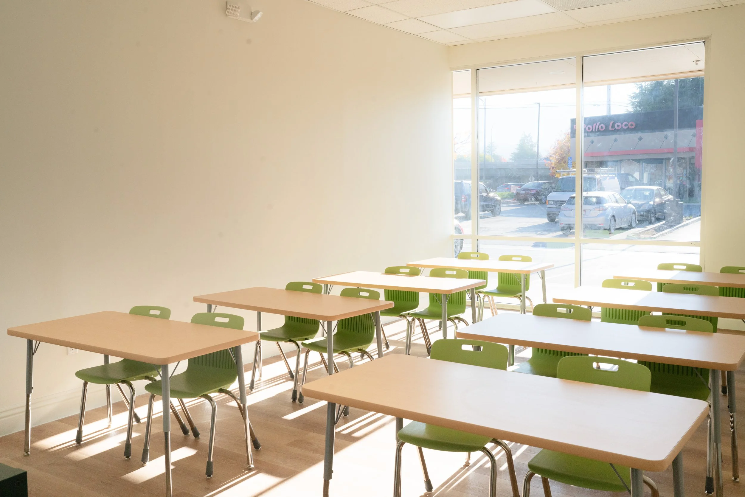 Empty classroom with green chairs and beige desks, large window showing parked cars outside, sunlight streaming in.