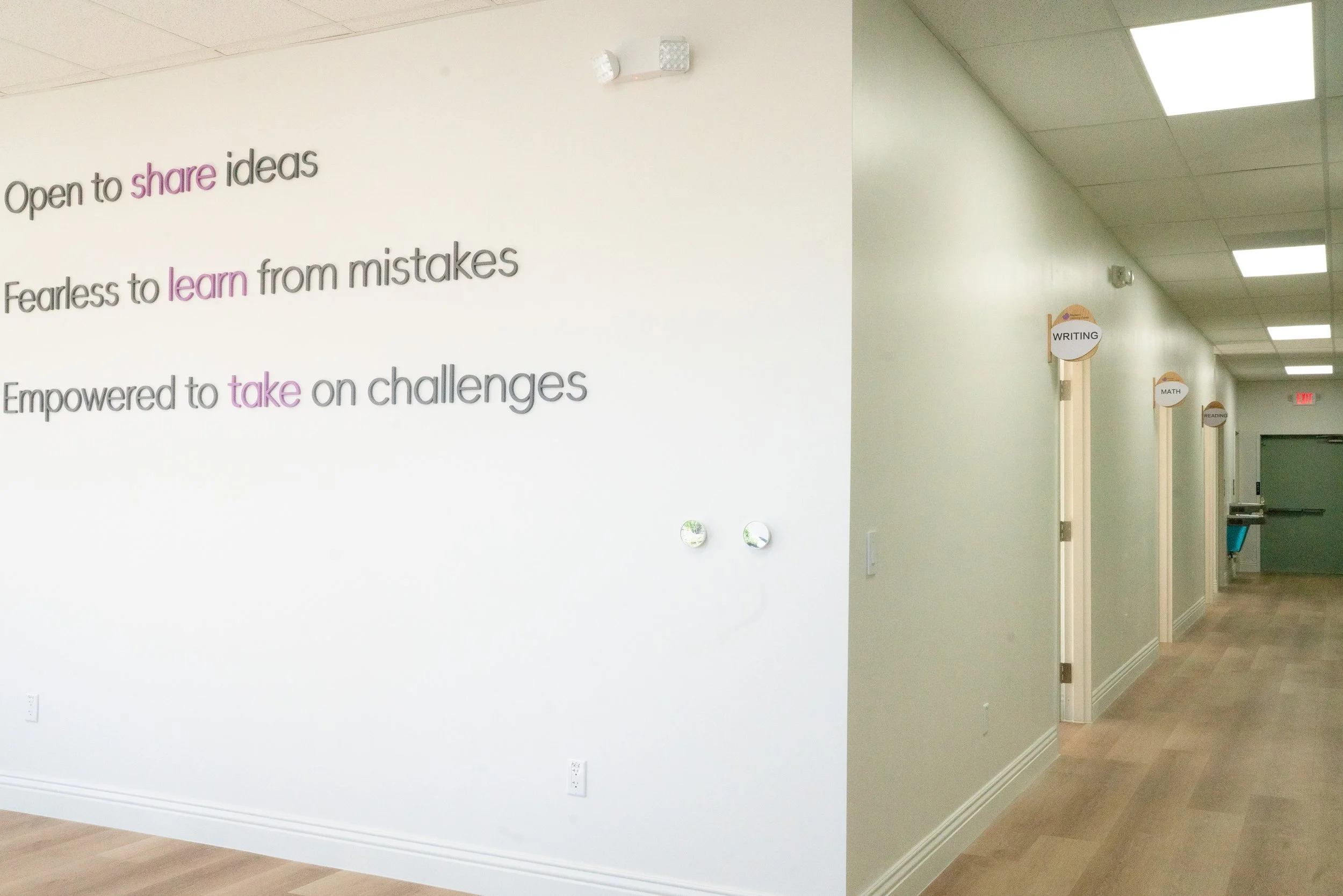 Hallway in a school or educational facility with signs indicating classrooms for writing, math, and reading, and a large wall with motivational quotes about sharing ideas, learning from mistakes, and taking on challenges.