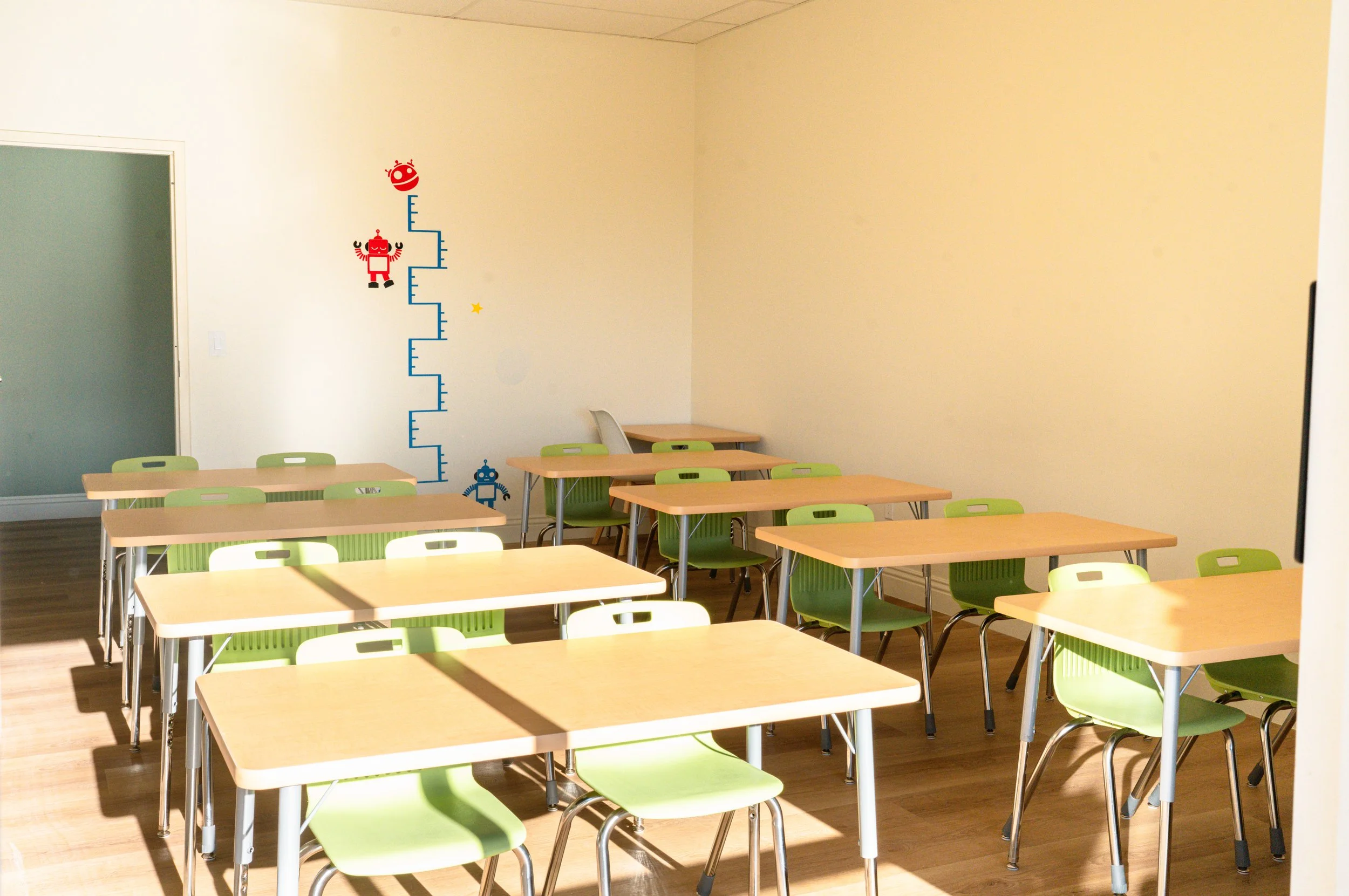 Classroom with wooden tables and green chairs, wall with height chart and robot decals, sunlight casting shadows.
