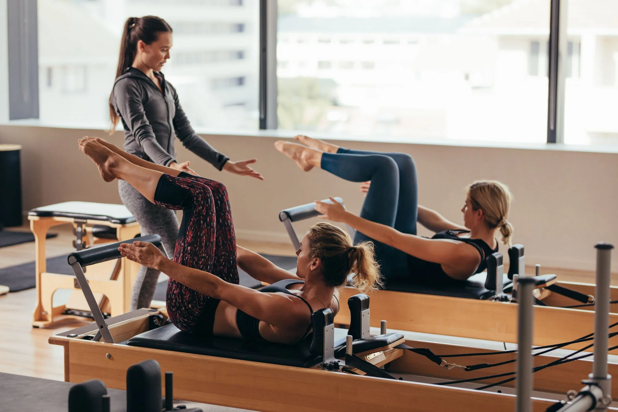 A Pilates instructor assisting women in performing exercises on reformer machines in a bright studio with large windows.