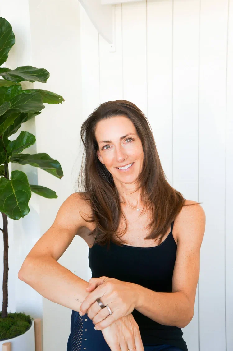 Smiling woman with long brown hair wearing a black tank top, standing indoors near green houseplant, with white wall background.