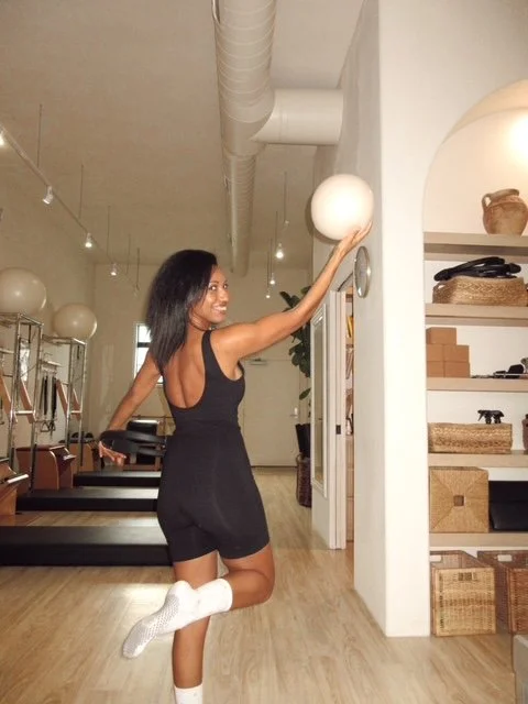 A woman in a black dress and white socks is jumping with one foot lifted behind her, reaching up to touch a white decorative orb on the wall, in a modern room with wooden floors and shelves.