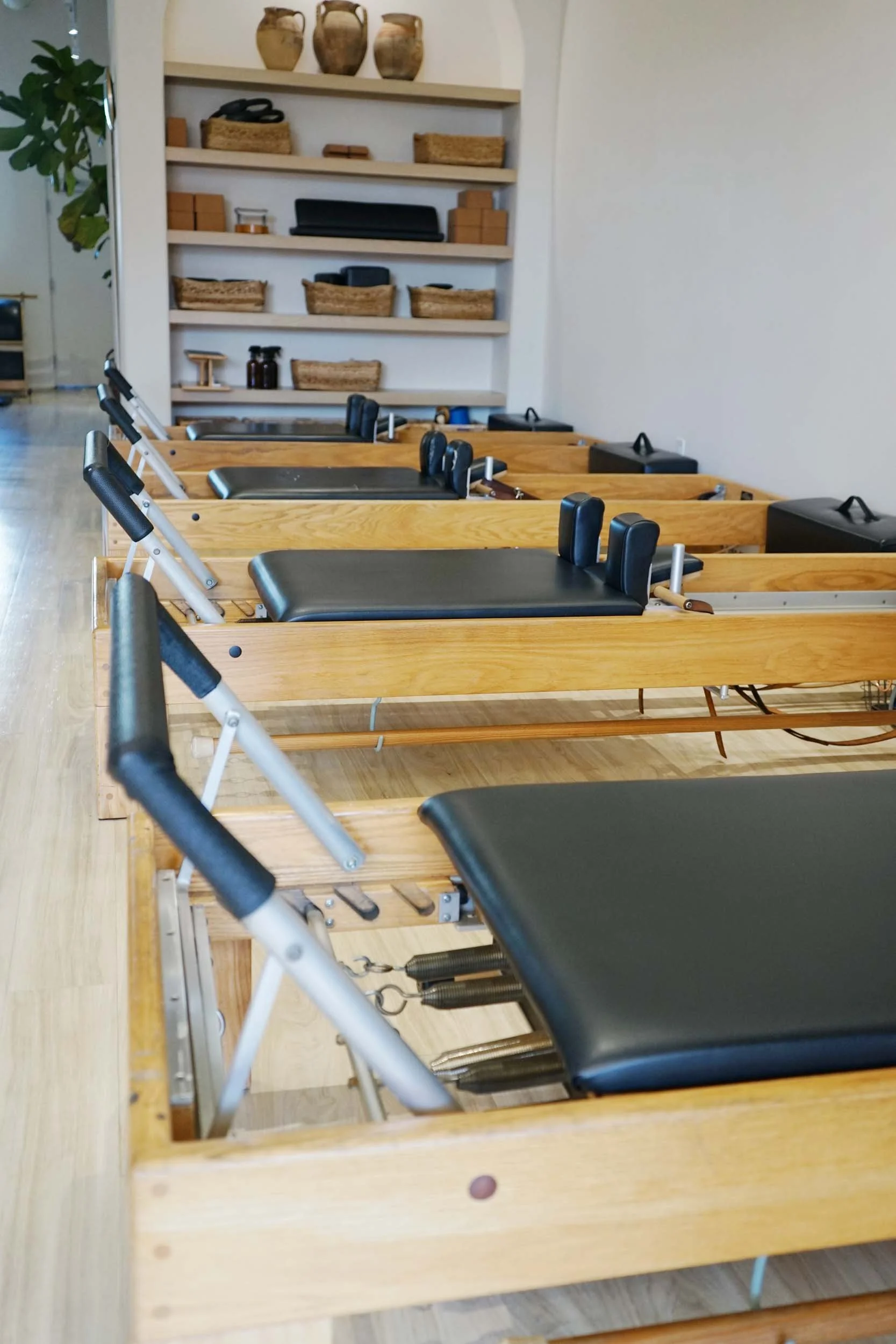 Chiropractic treatment tables arranged in a row in a wellness clinic, with wooden frames and black padding, against a background of shelves with pottery and baskets.