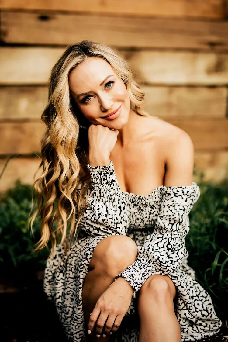 A woman with long blonde wavy hair, smiling, sitting outdoors in front of a wooden fence, wearing an off-shoulder black and white patterned dress.