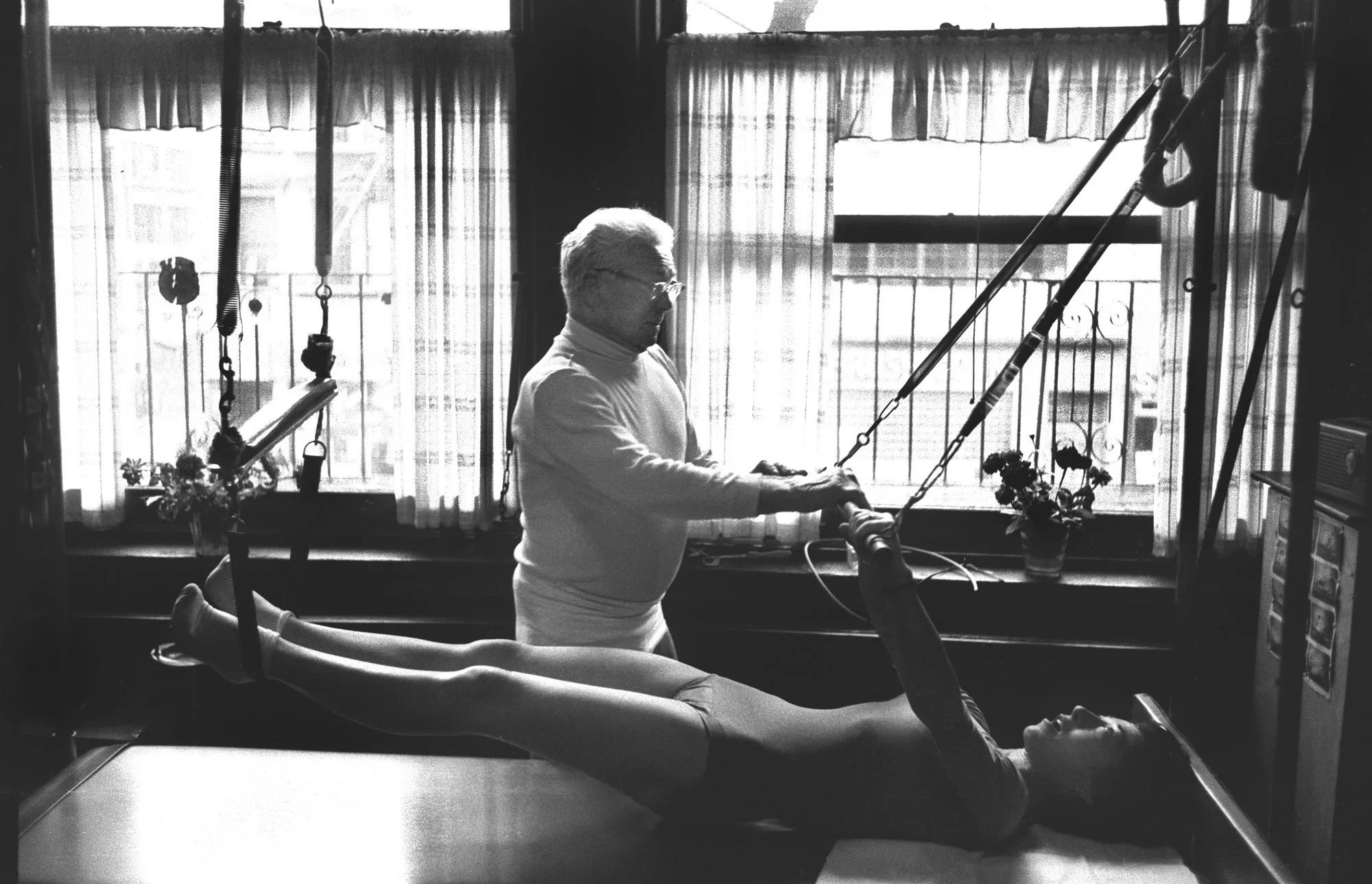 A person lying on a hospital bed with an IV drip in their arm, being examined or treated by an elderly man in a room with large windows and curtains.