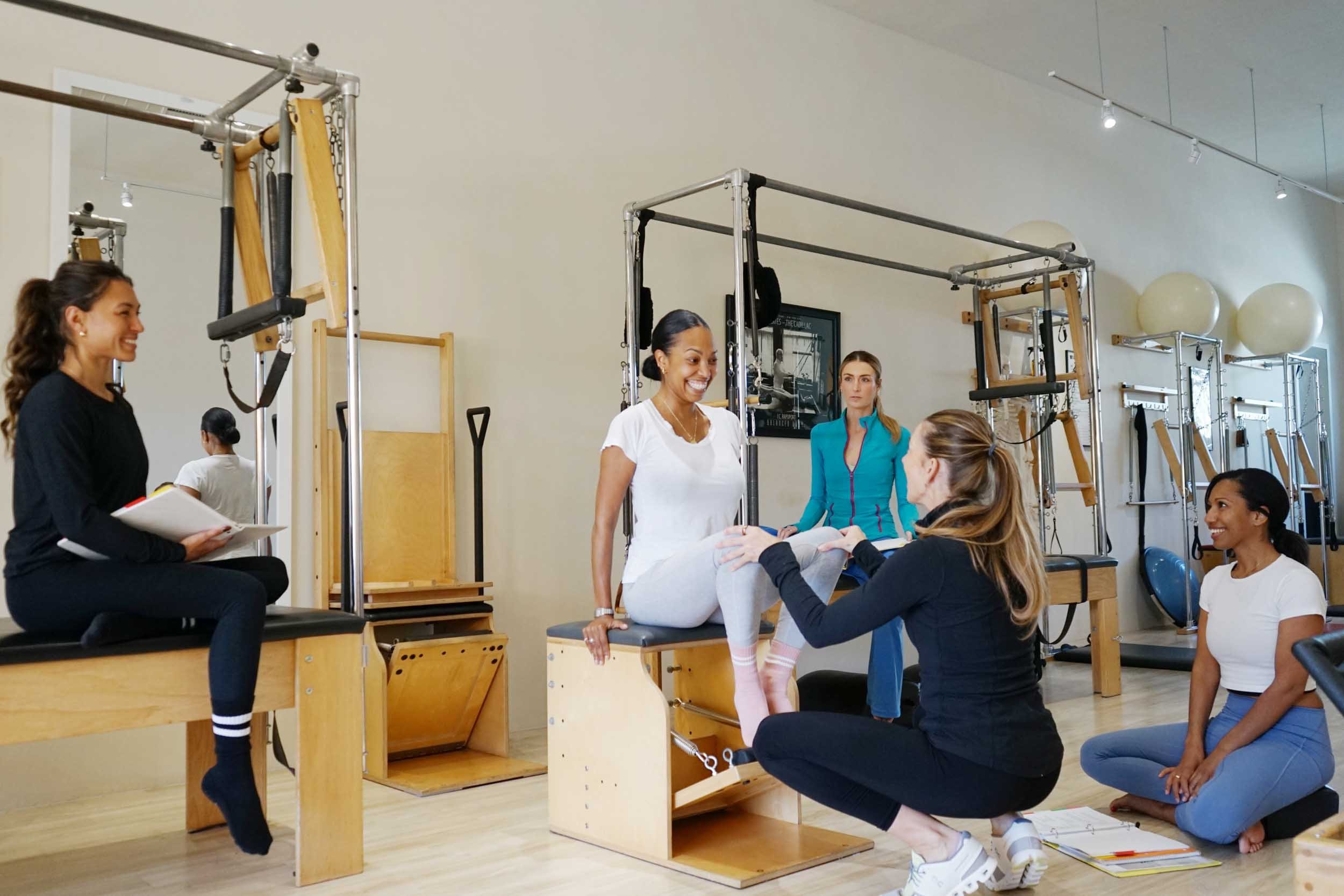 Group of women in a physical therapy or Pilates studio, with one woman sitting on a wooden box and others assisting or observing, surrounded by Pilates equipment and exercise balls.