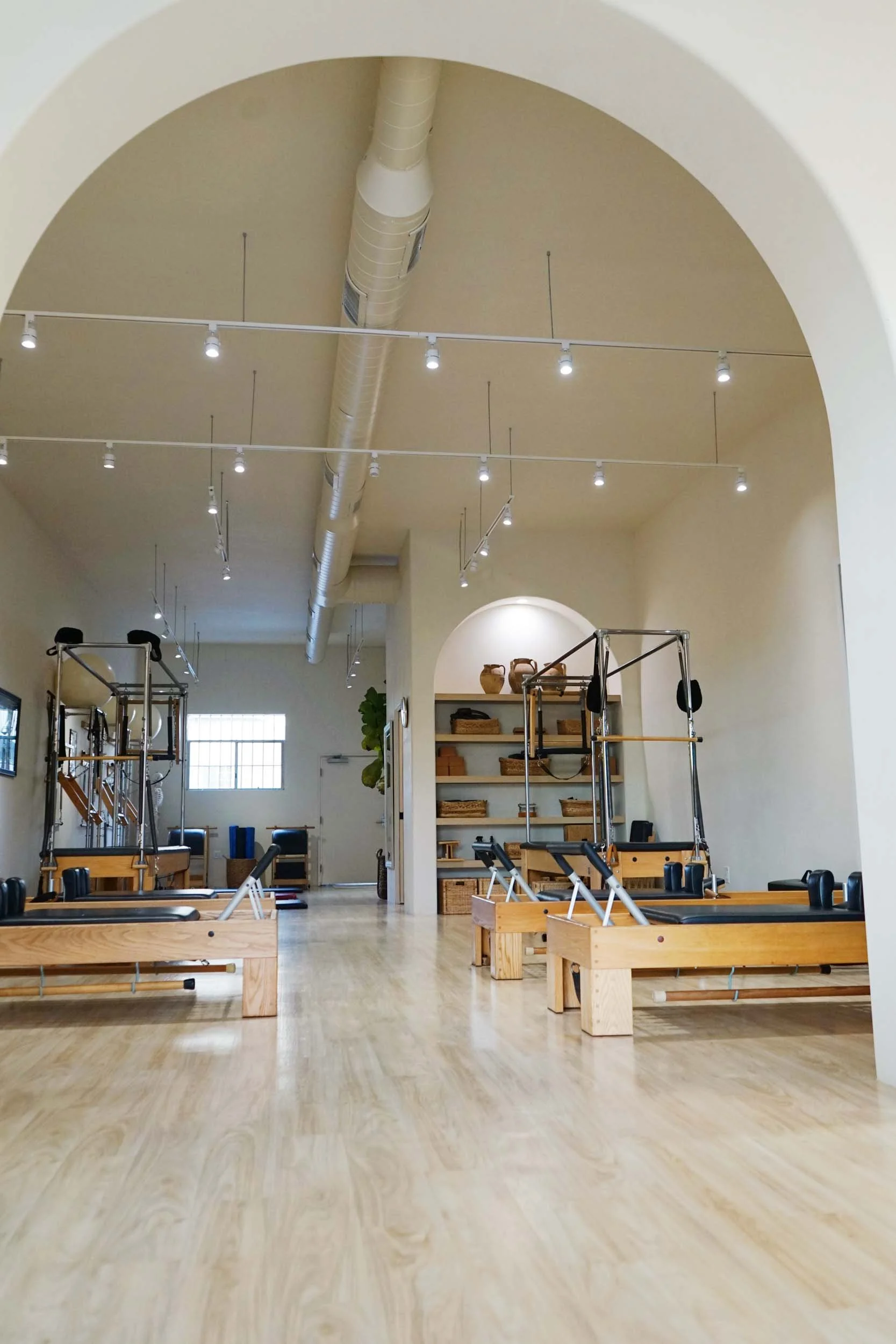 An open Pilates studio with wooden reformer equipment, shelves with baskets, and warm lighting. High ceiling with visible ductwork.