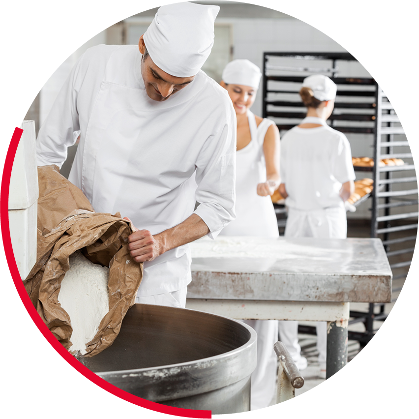 Trois pâtissiers en uniforme travaillant dans une boulangerie, avec des étagères de pains en arrière-plan.