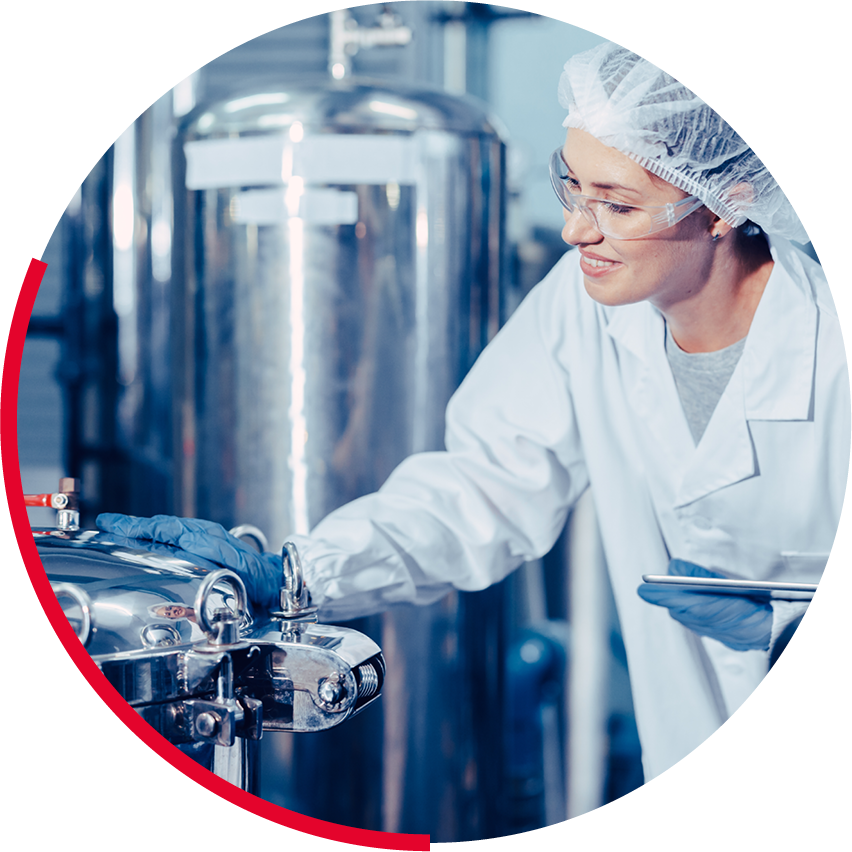 Une femme en blouse de laboratoire, portant des lunettes de protection, examine une machine industrielle dans un laboratoire ou une usine.