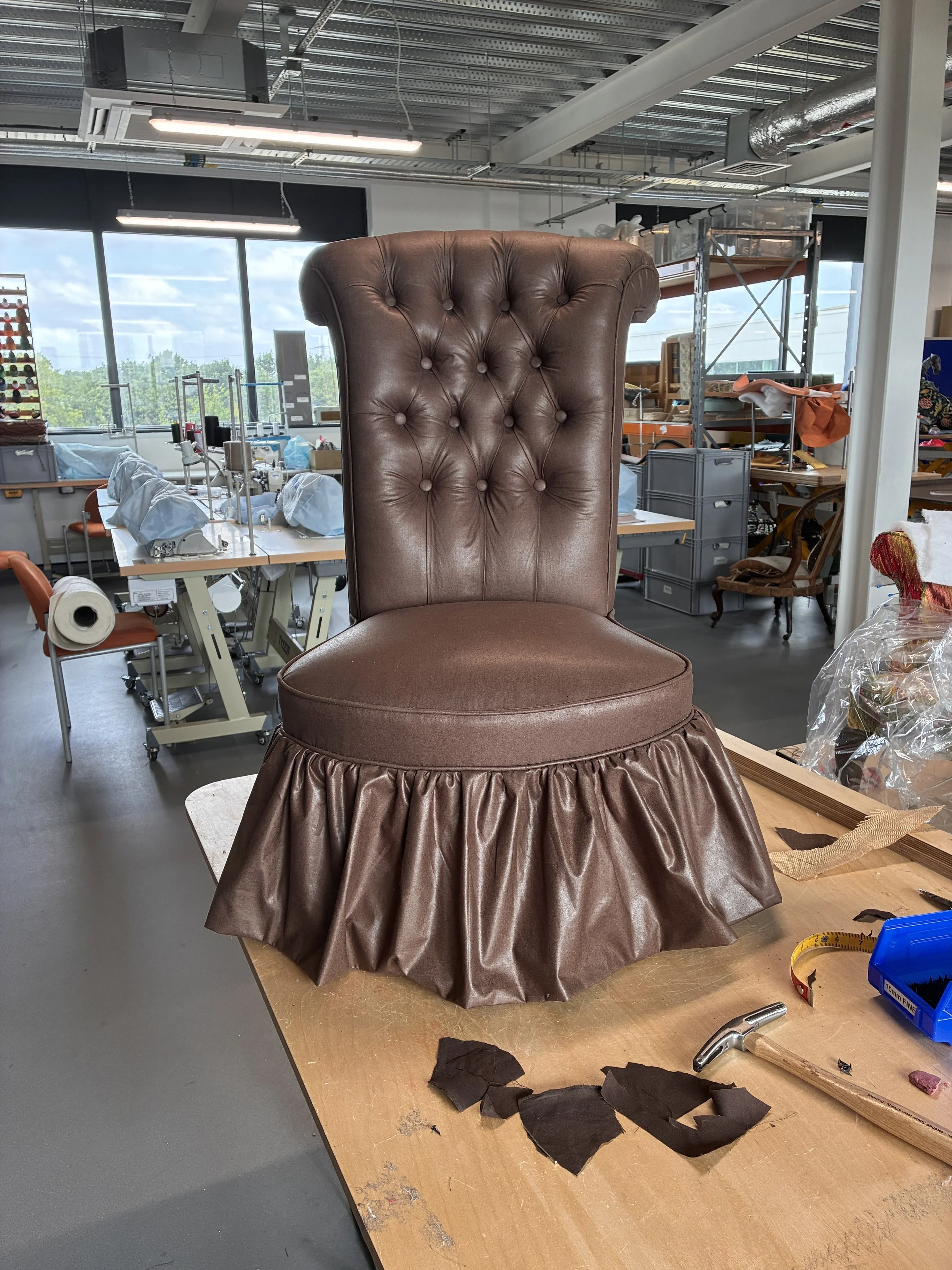 A custom upholstered chair with a tall, tufted brown leather backrest and a gathered brown fabric skirt at the base, in a workshop with sewing and upholstery materials around.