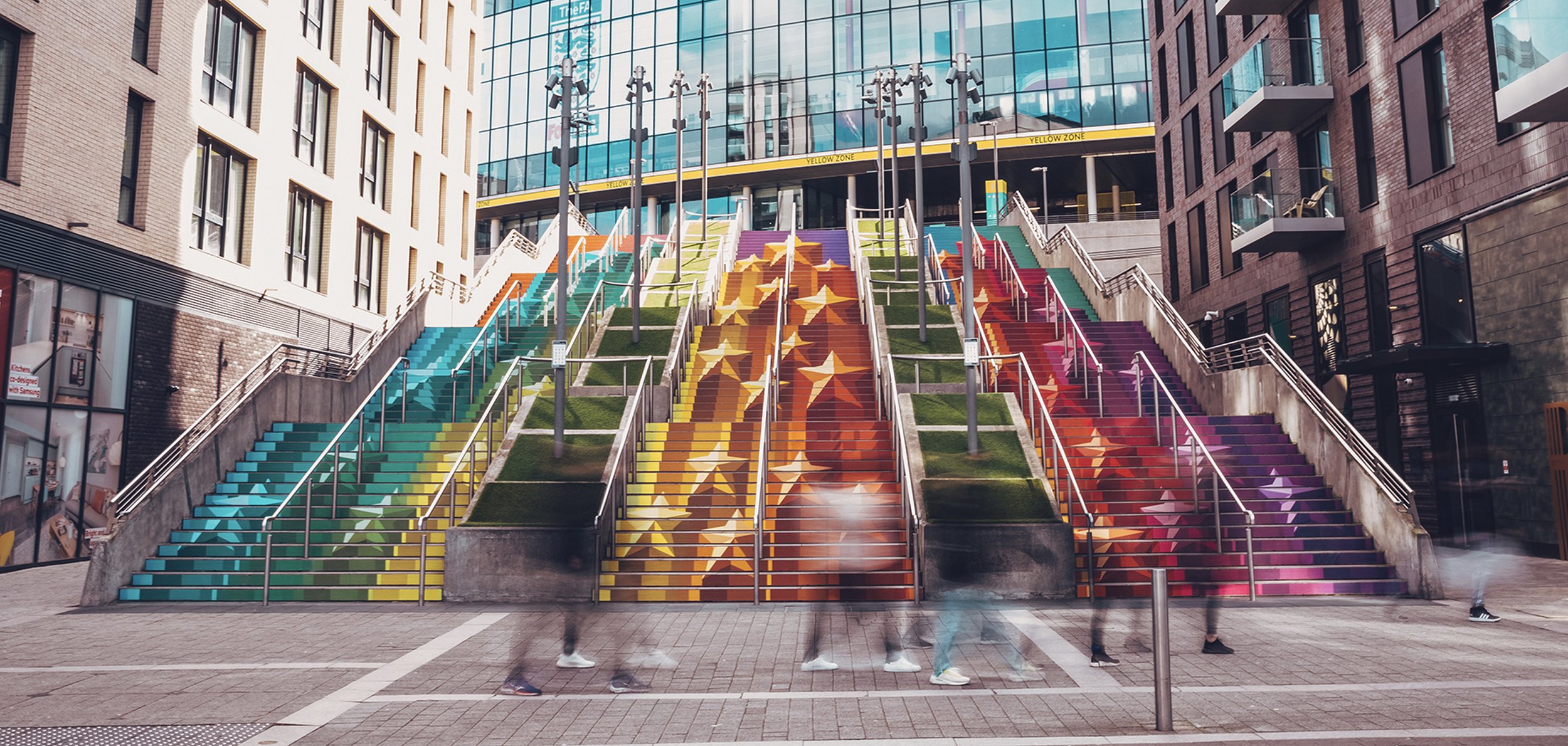 Colorful outdoor staircase with rainbow-colored steps, leading to a modern glass building, with blurred people walking in front.