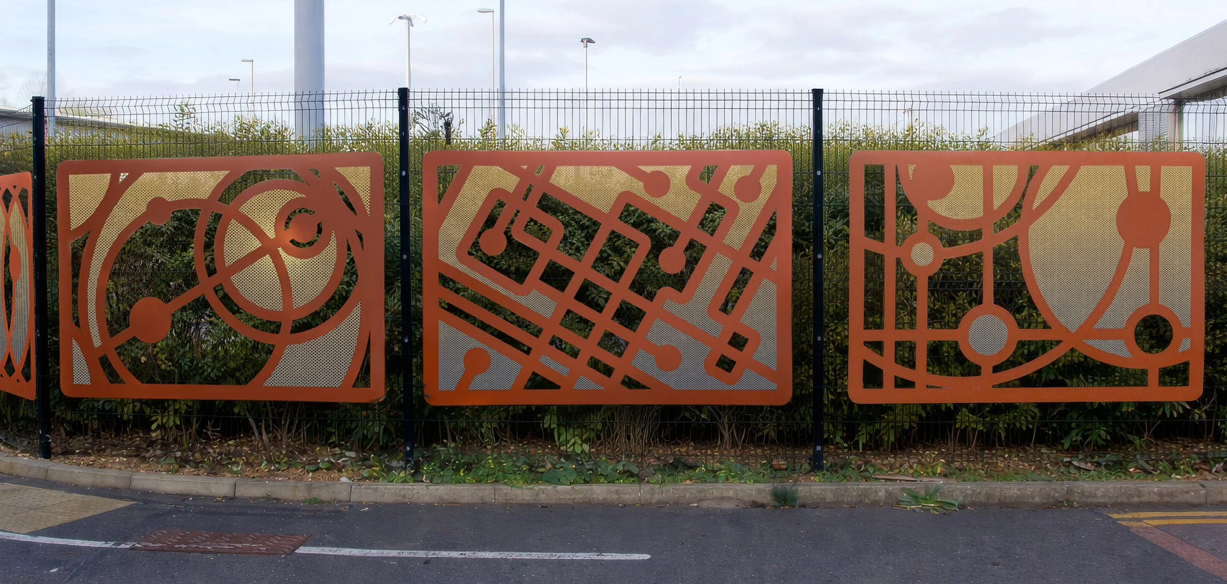 Three orange metal panels with abstract geometric cutout designs, placed in front of a green hedge, with a paved sidewalk and overcast sky in the background.