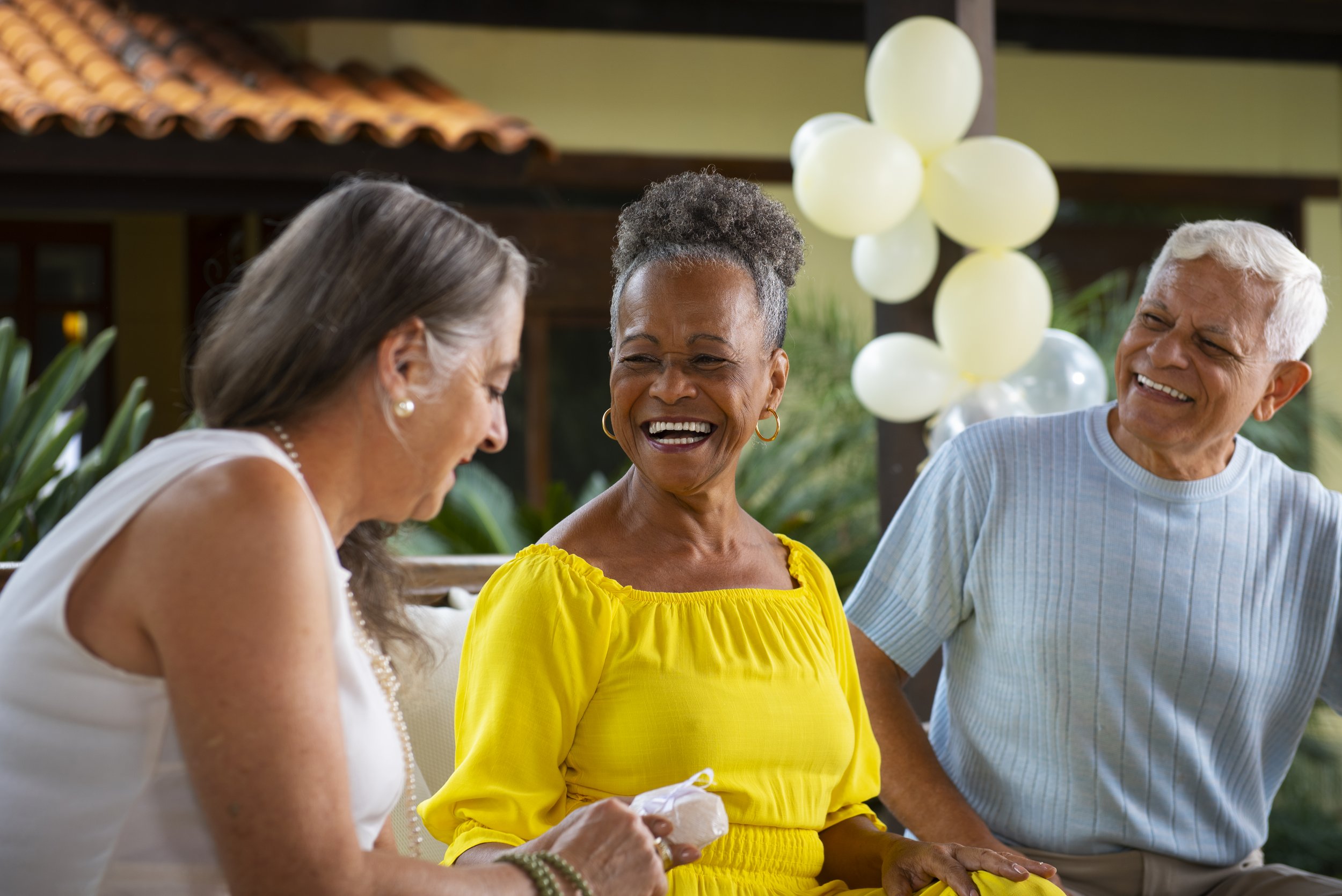 Three older adults sitting outdoors, smiling and laughing, at a gathering with balloons in the background.