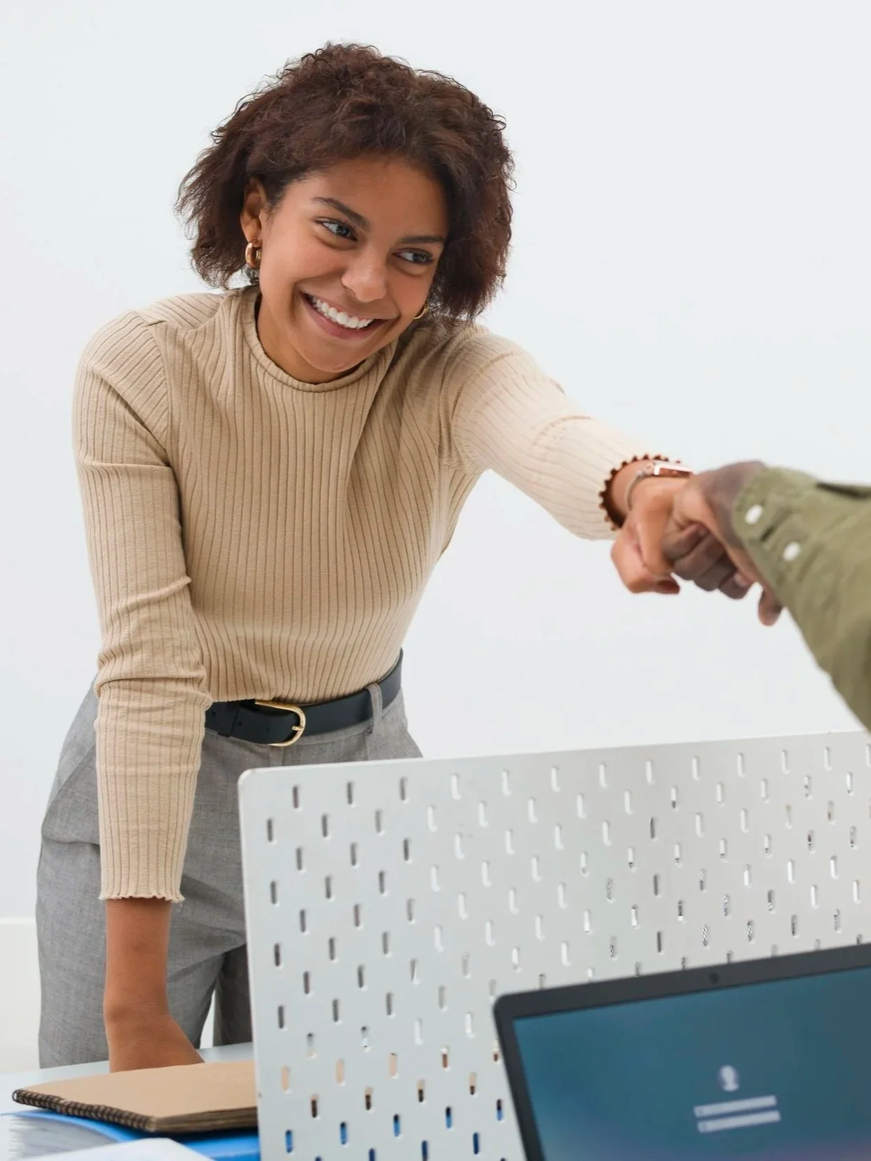 Smiling woman in beige ribbed sweater and gray pants shaking hands in an office setting.